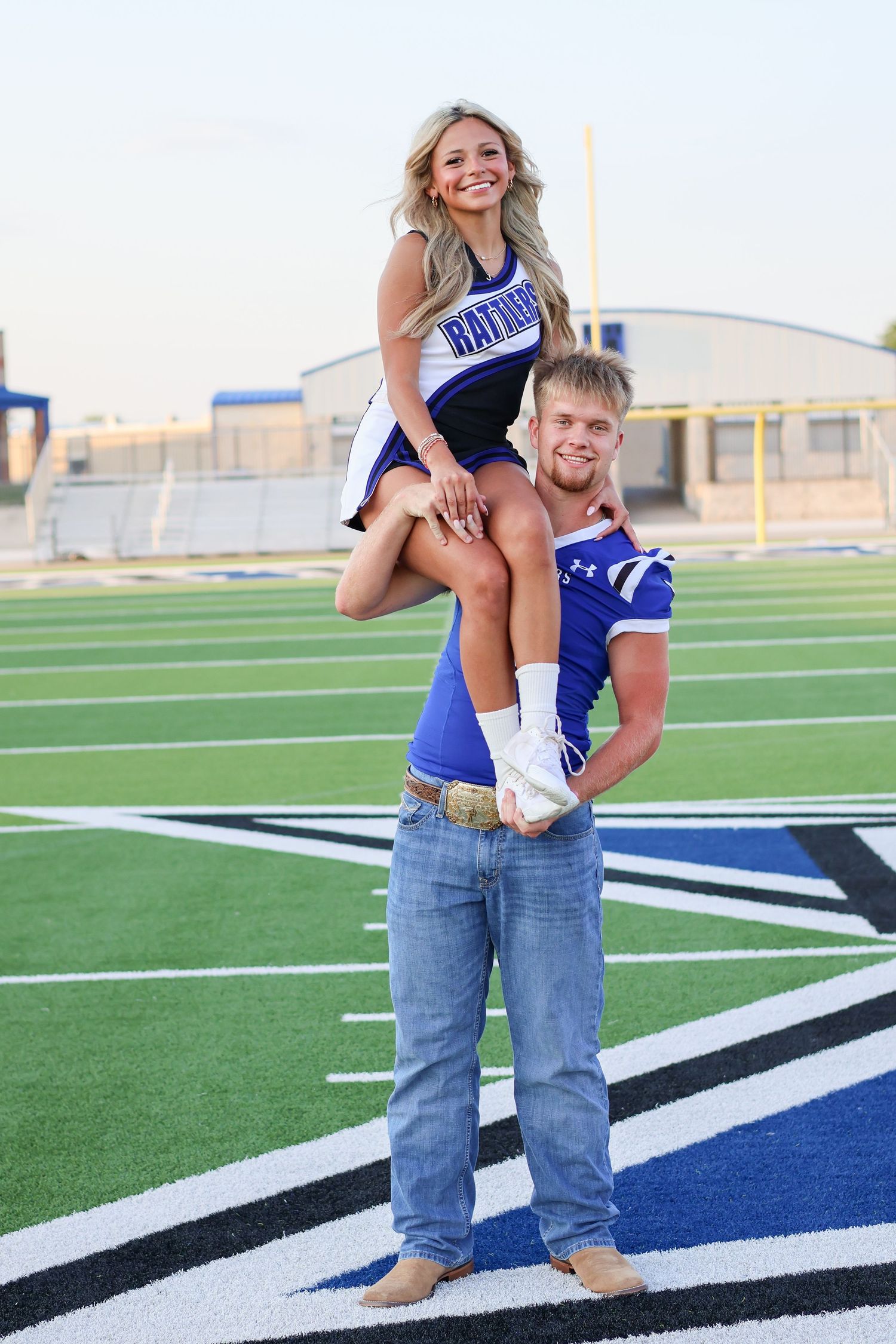 Cheerleader being lifted by football player on field with stadium bleachers in background.