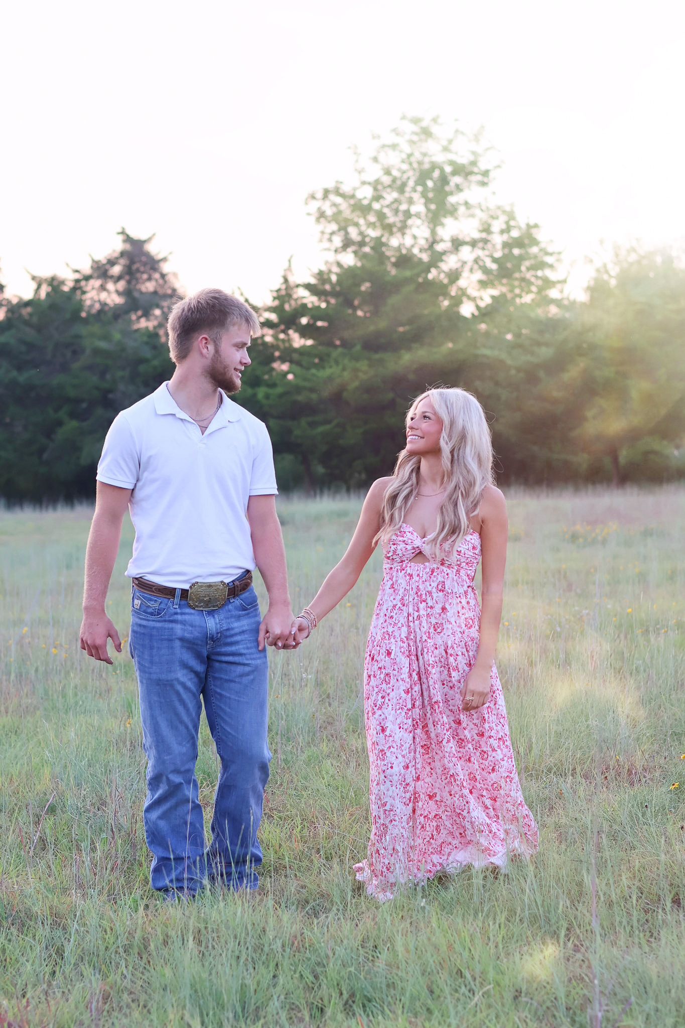 A couple holds hands while walking through a sunlit meadow at golden hour, wearing casual summer attire.