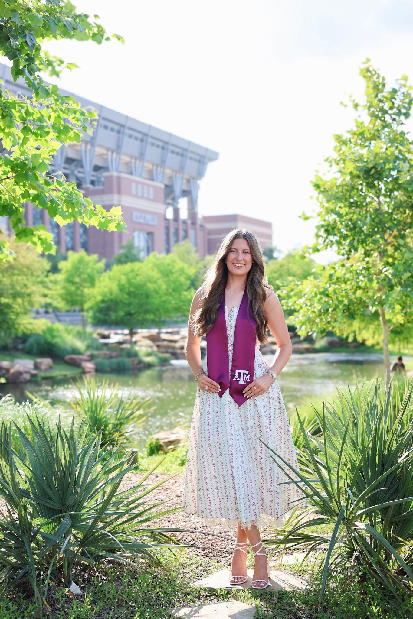Young woman in white dress and maroon Texas A&M graduation stole smiling outdoors near a pond on campus.