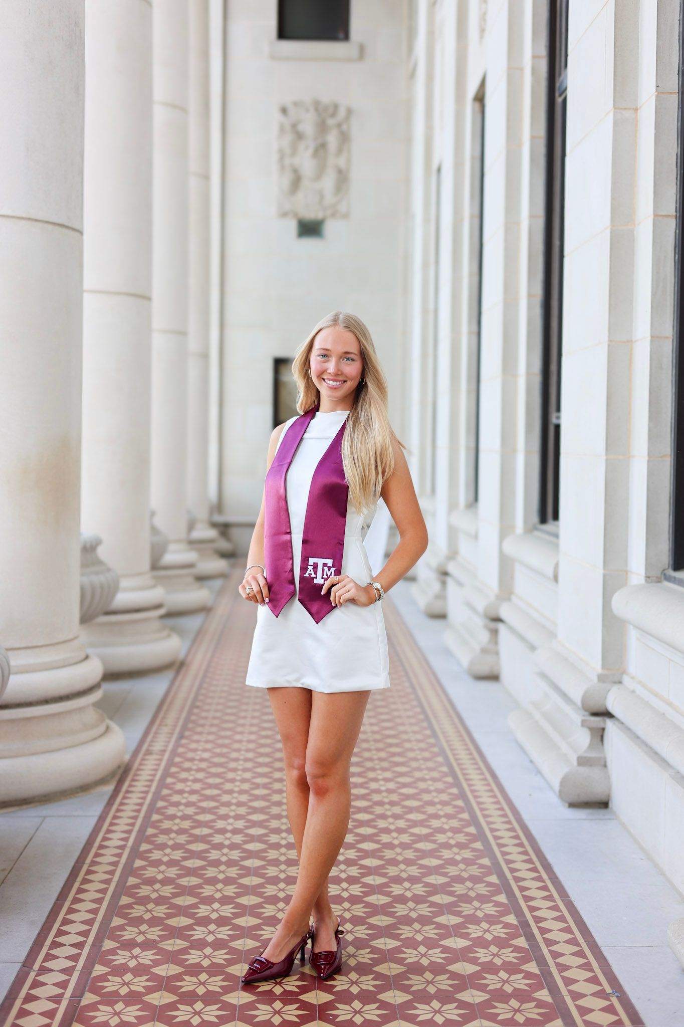 Texas A&M graduate smiling in white dress with maroon graduation stole, posing between elegant columns on campus.