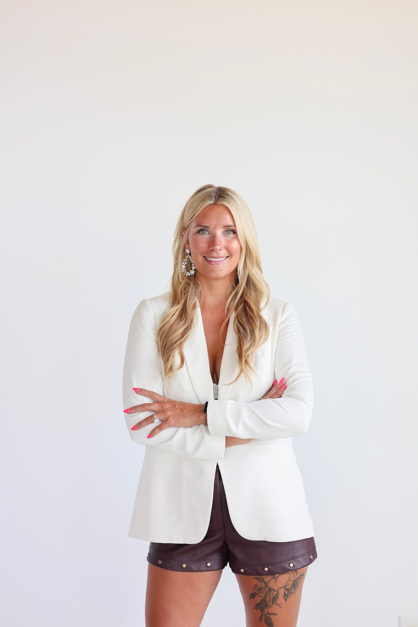 Smiling blonde woman in white blazer and brown shorts stands with arms crossed against white background.