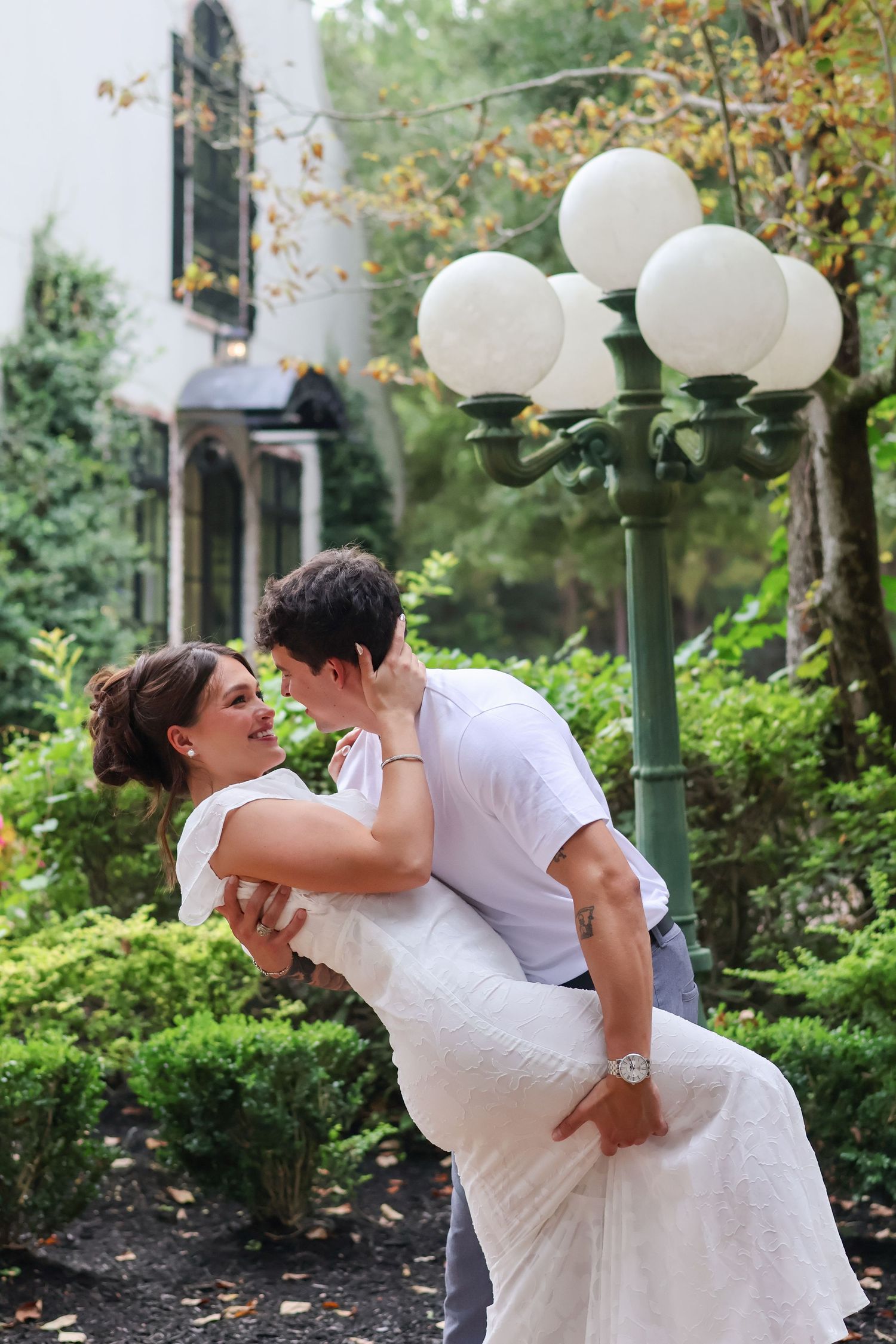 A romantic couple in wedding attire dances and embraces near a decorative outdoor lamp post in a garden setting.