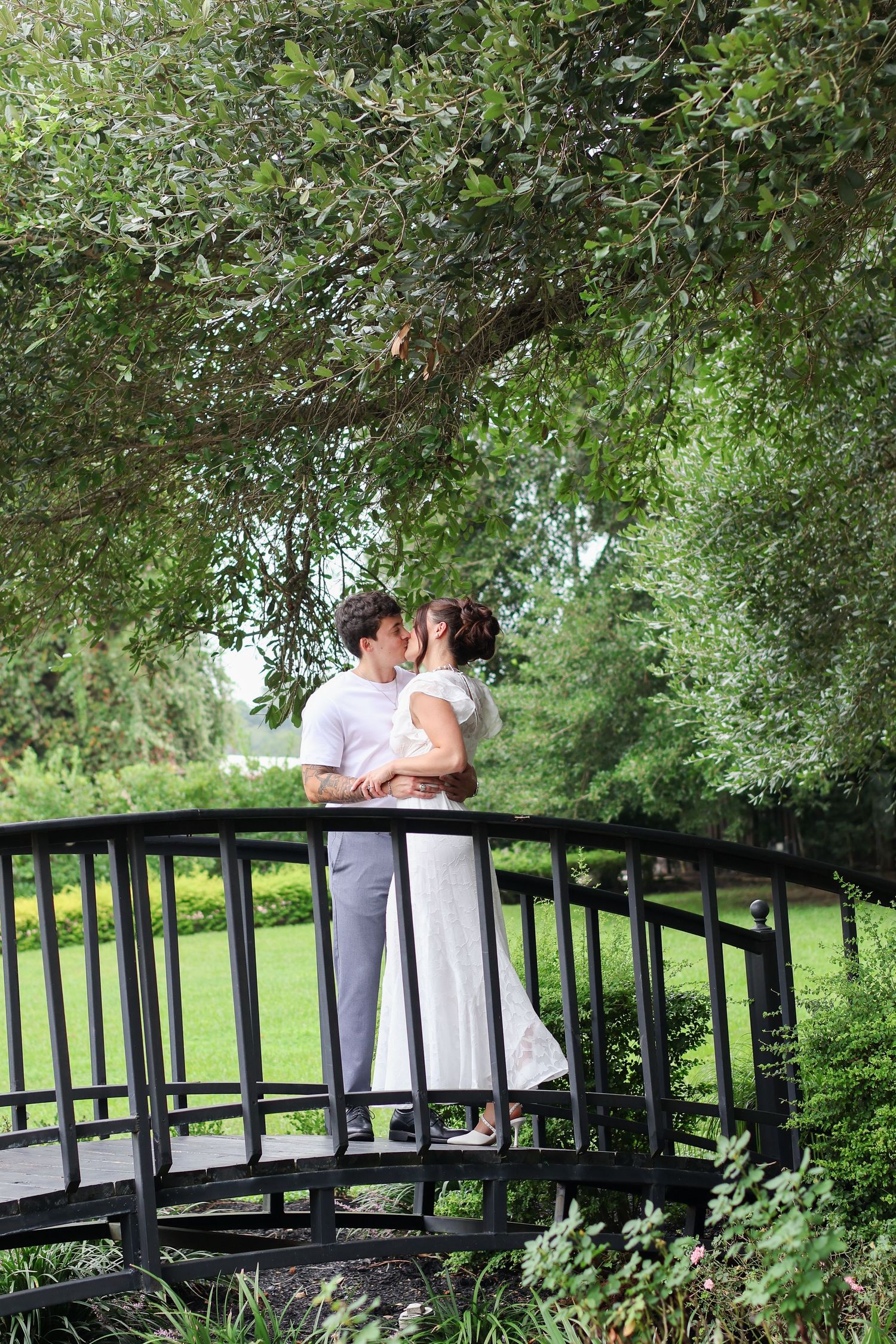 A couple shares a romantic moment on a wooden footbridge surrounded by lush green trees in a garden setting.