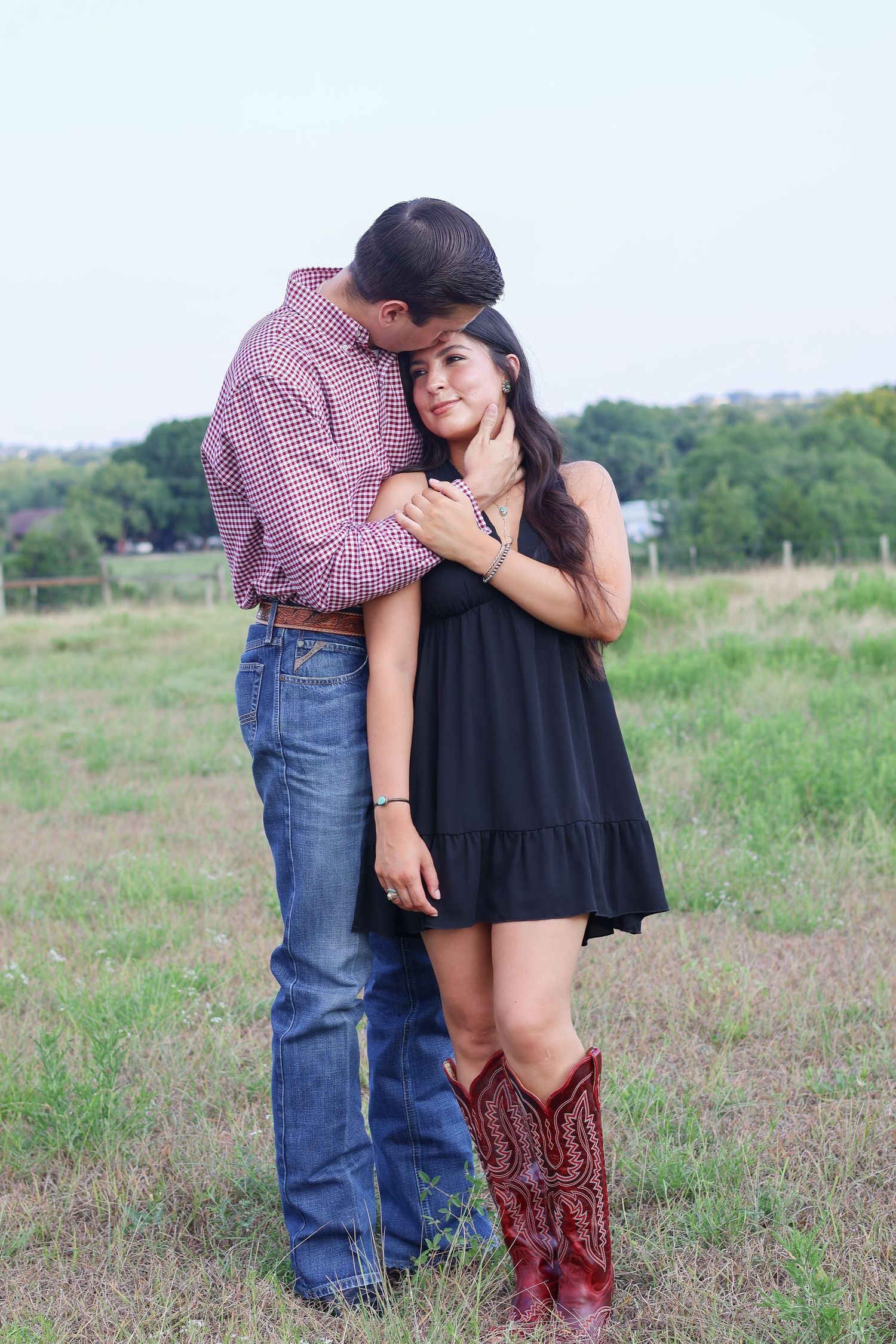 A romantic couple embraces in a grassy field while wearing western attire including cowboy boots and a baseball cap.