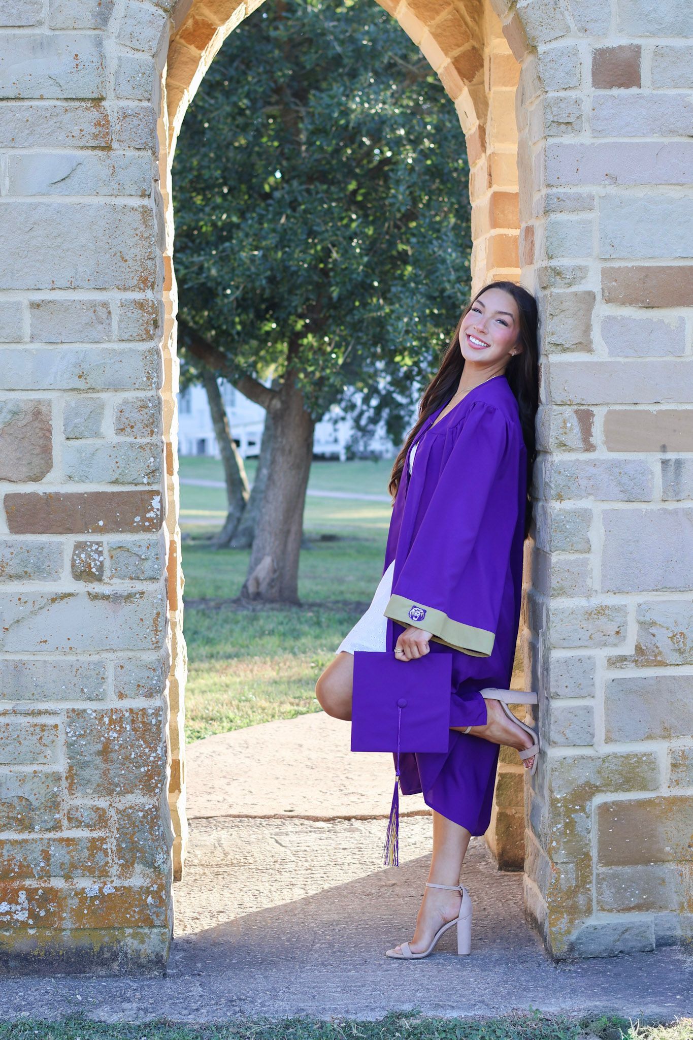 Smiling graduate in purple cap and gown poses in stone archway outdoors on a sunny day.