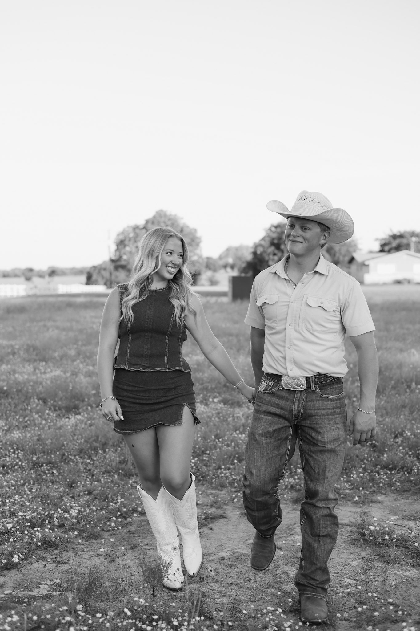 Black and white photo of a smiling couple holding hands walking through a field, woman in mini dress and boots, man in cowboy hat.