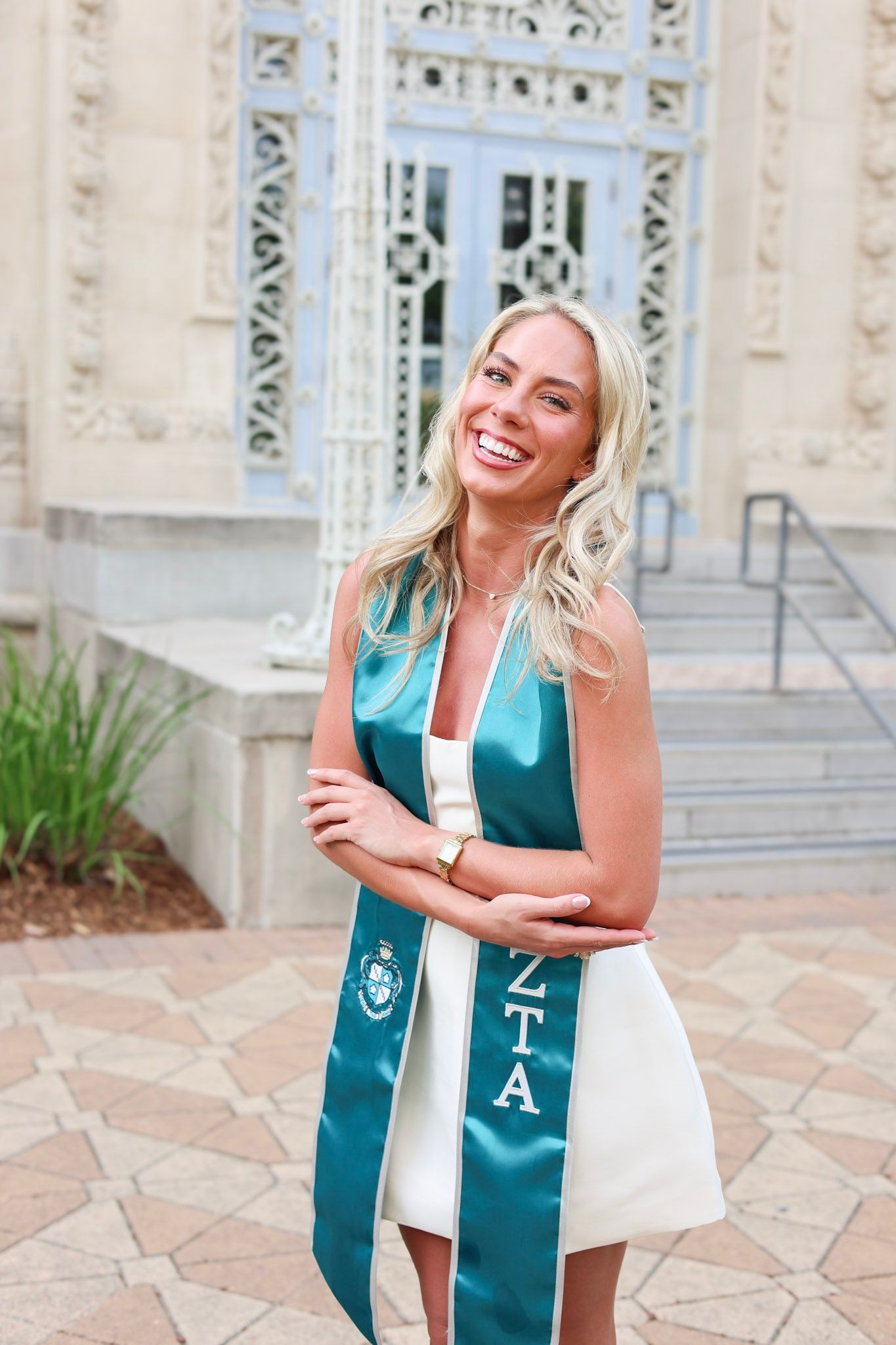 Smiling blonde woman wearing a teal Zeta Tau Alpha graduation sash poses in front of a university building.