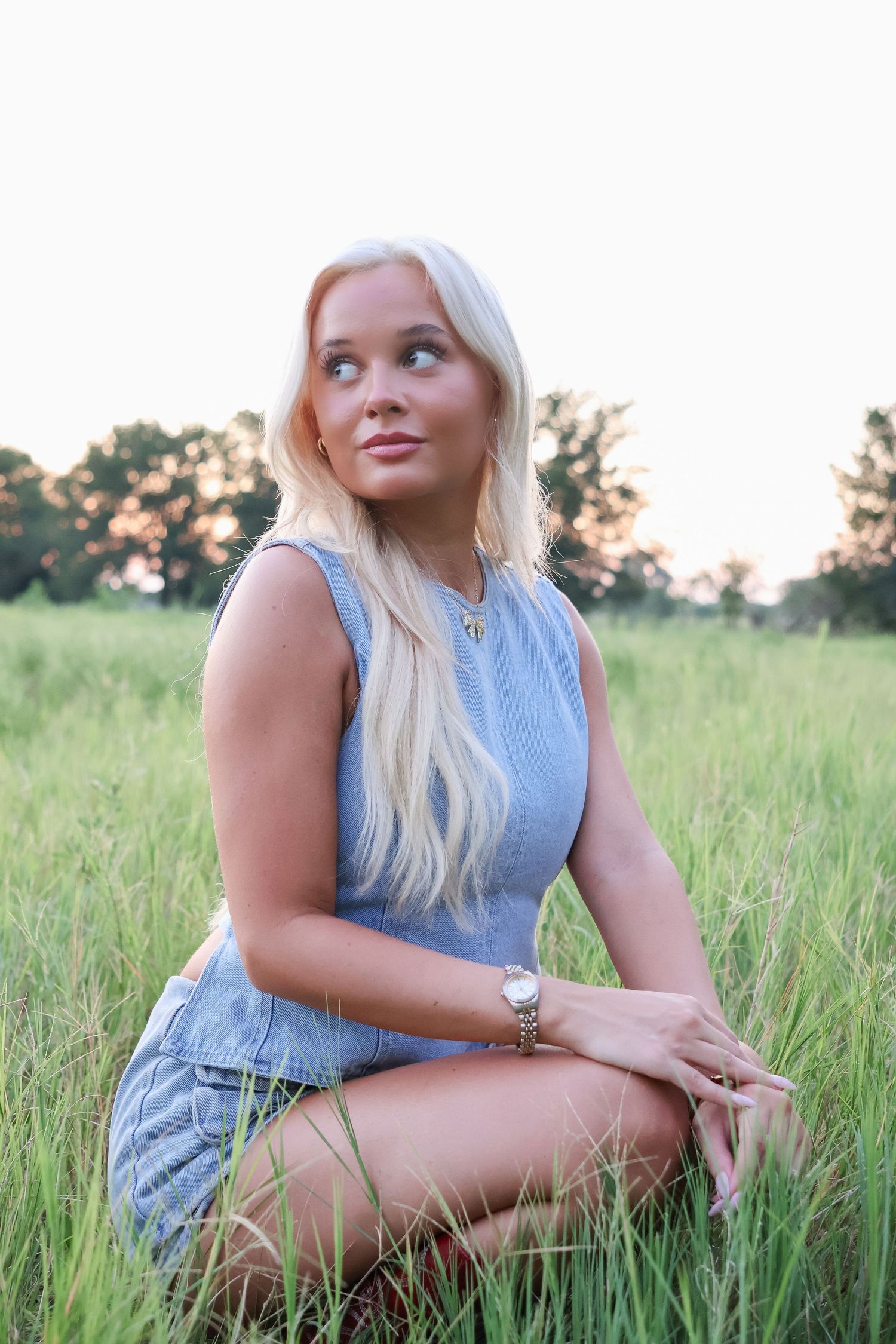 Someone poses in a casual denim outfit while sitting in tall grass during sunset.