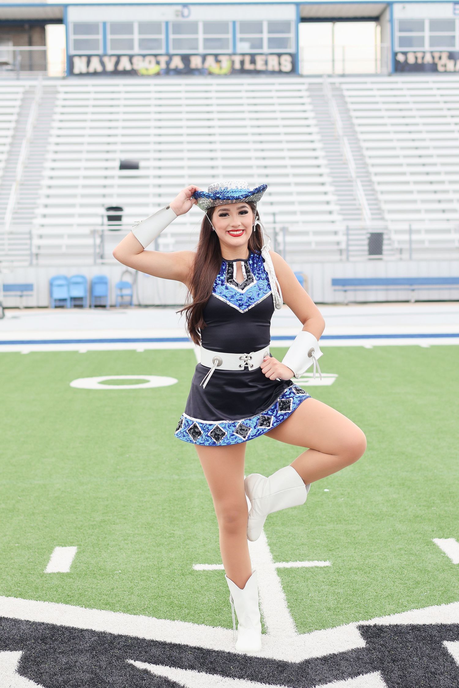 Cheerleader poses in blue and black uniform with white boots on football field in front of empty stadium bleachers.