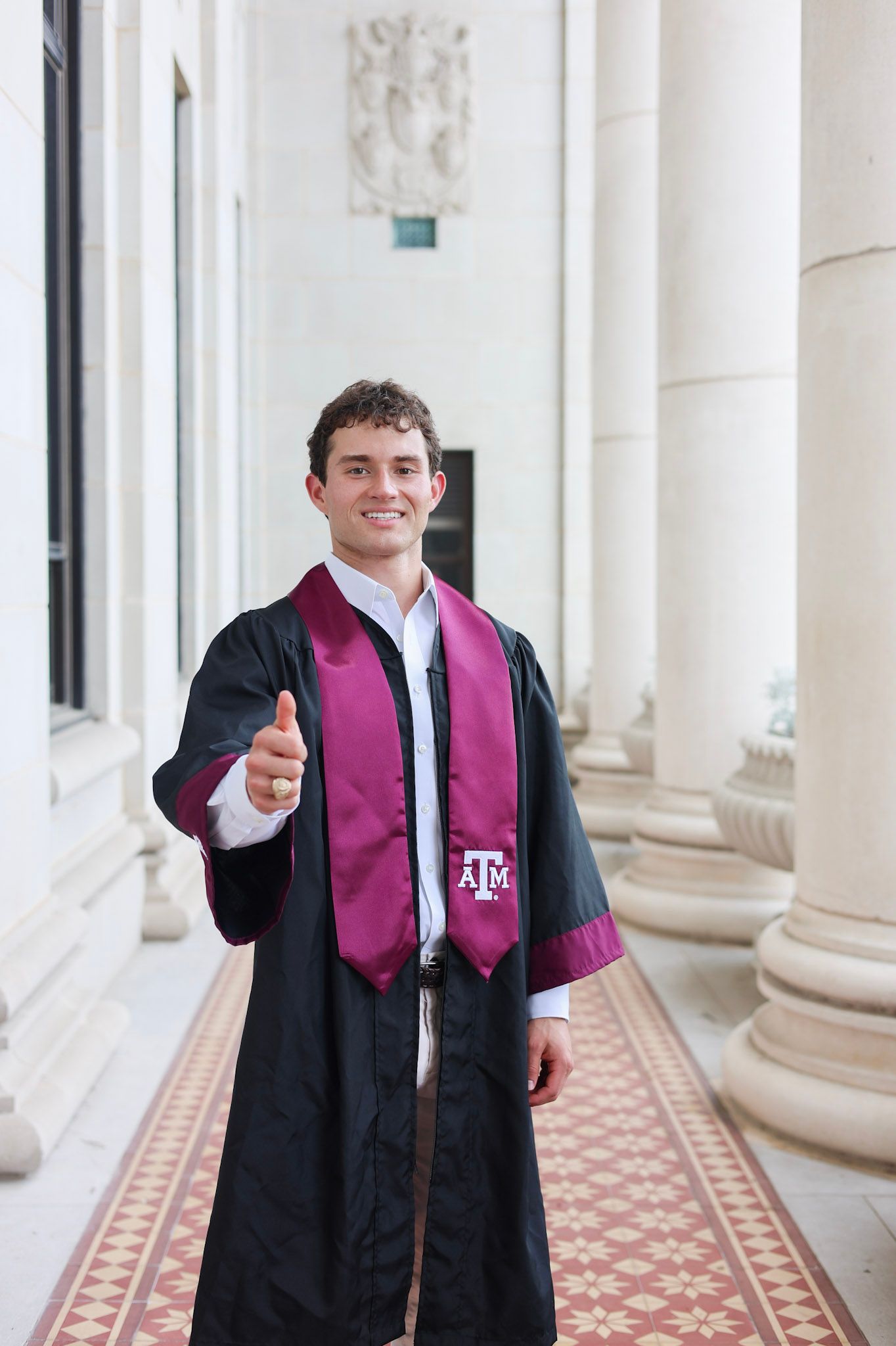 Texas A&M graduate in black robe and maroon stole gives thumbs up between columns.