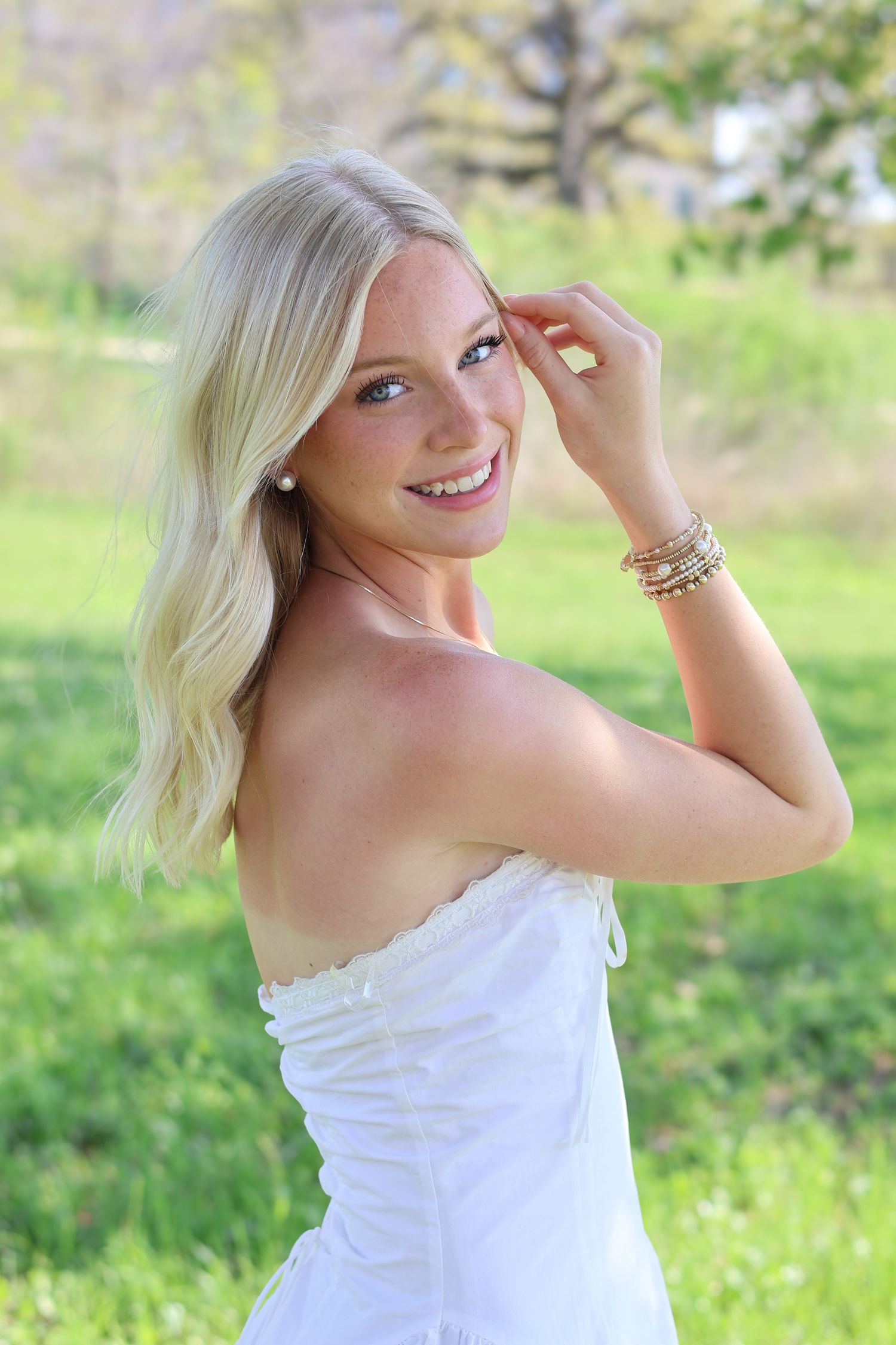 Smiling blonde woman in white strapless dress with gold bracelets posing outdoors in green park setting.