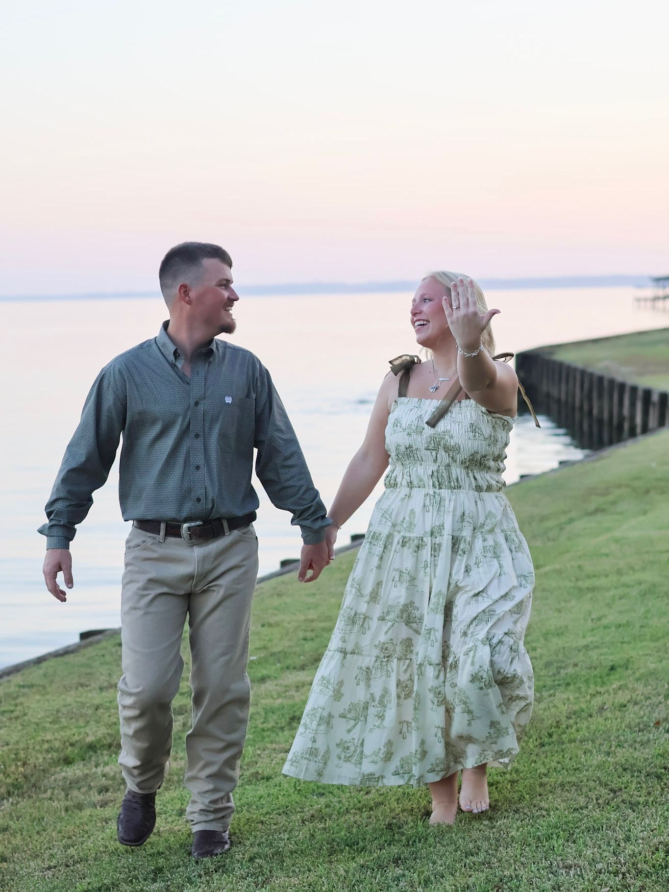 Couple walking hand in hand by waterfront at sunset, woman in floral dress waving, man in gray shirt and khakis.