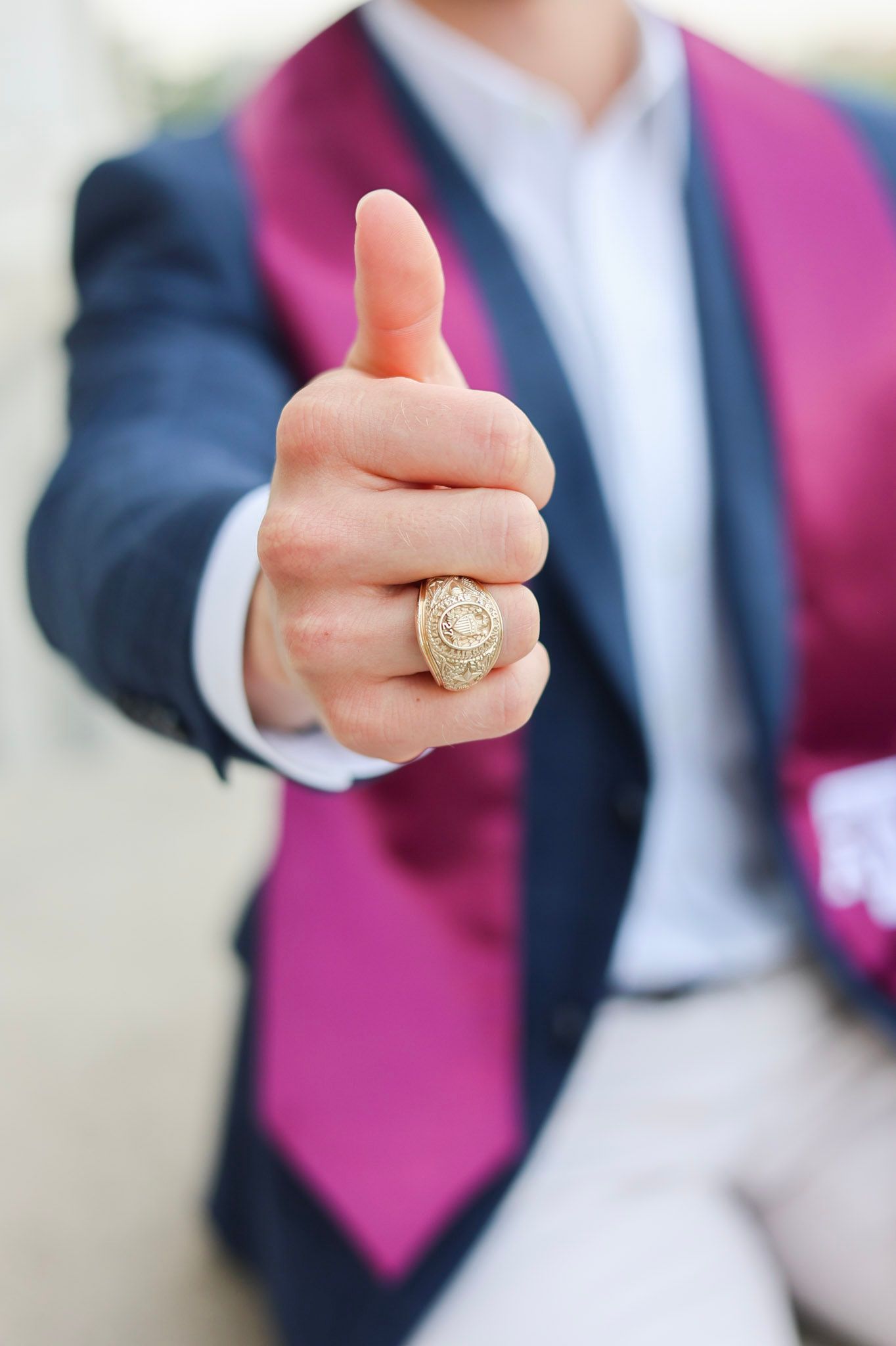 Graduate in blue blazer and pink sash gives thumbs up wearing gold class ring.