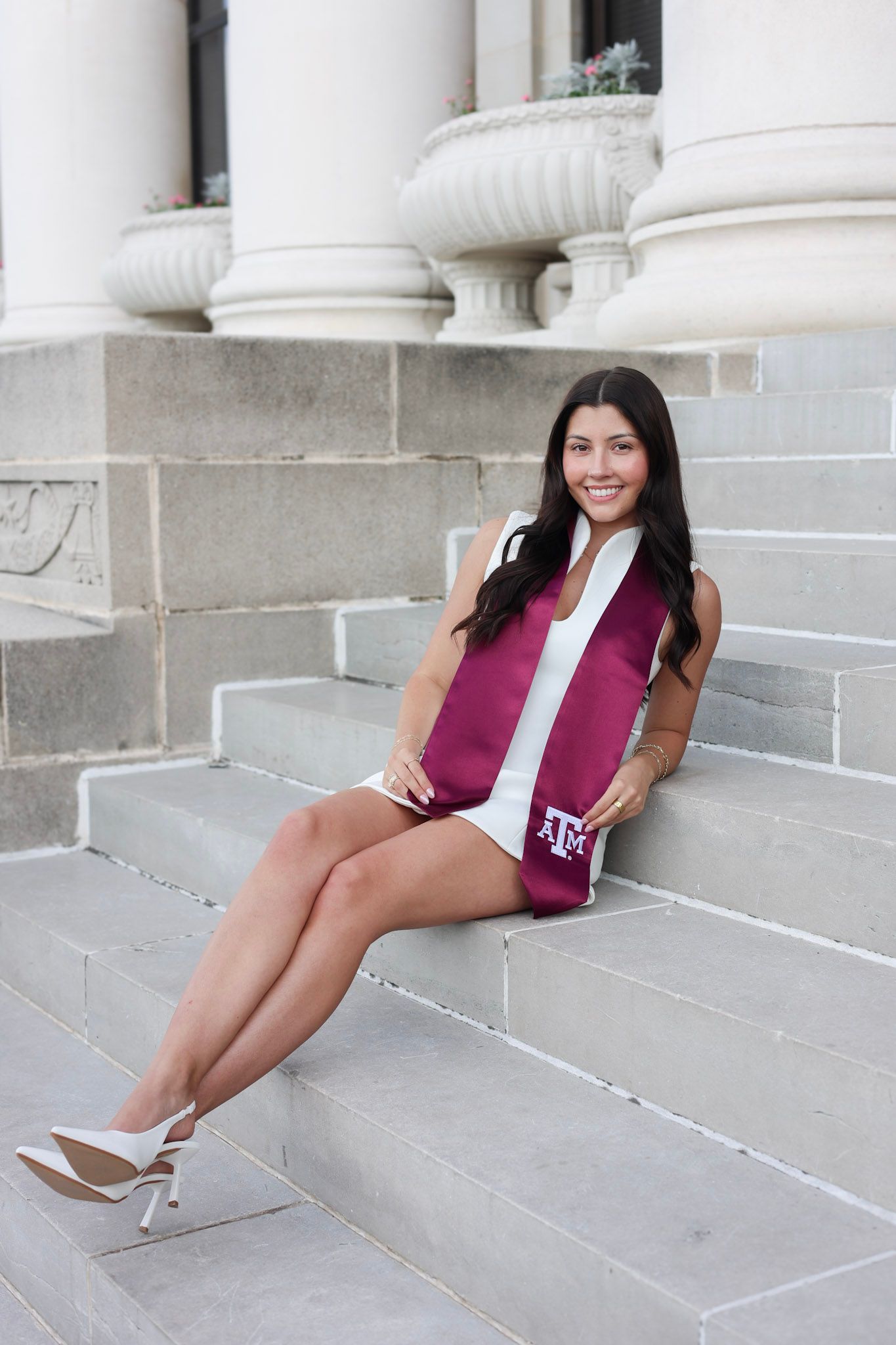 Young woman in white dress with Texas A&M maroon graduation stole sits smiling on stone steps.