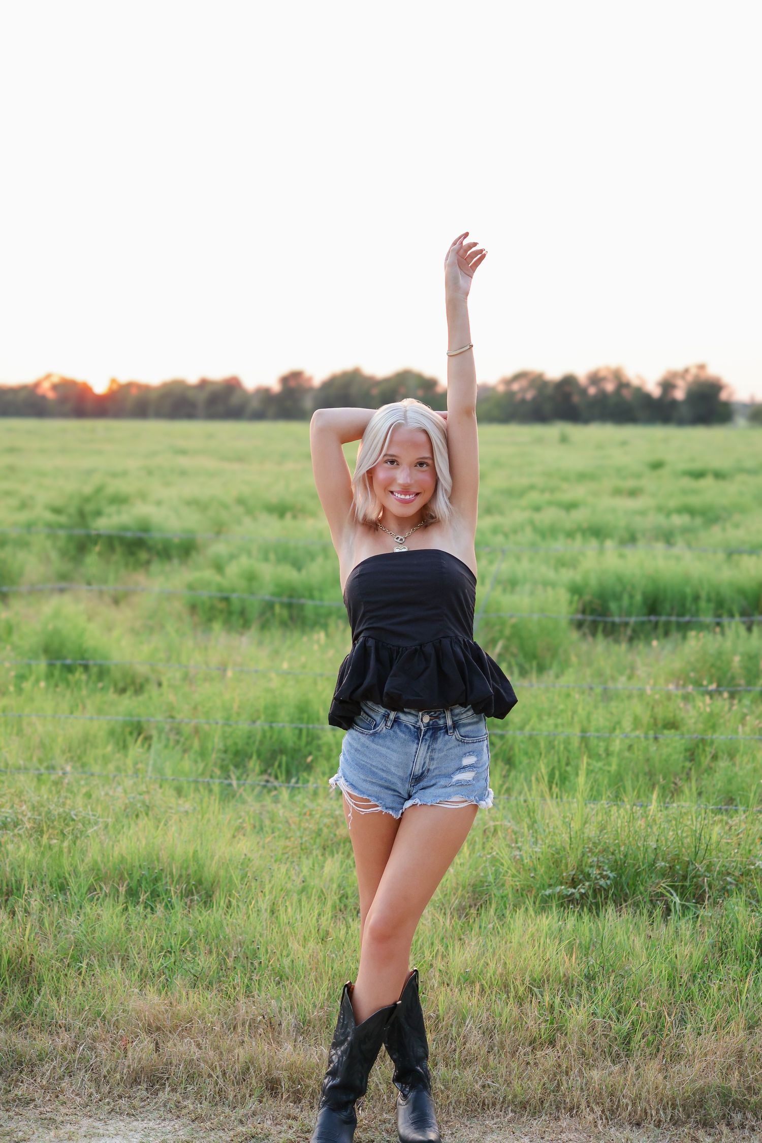 Woman wearing black top and denim shorts poses in a grassy field at sunset.