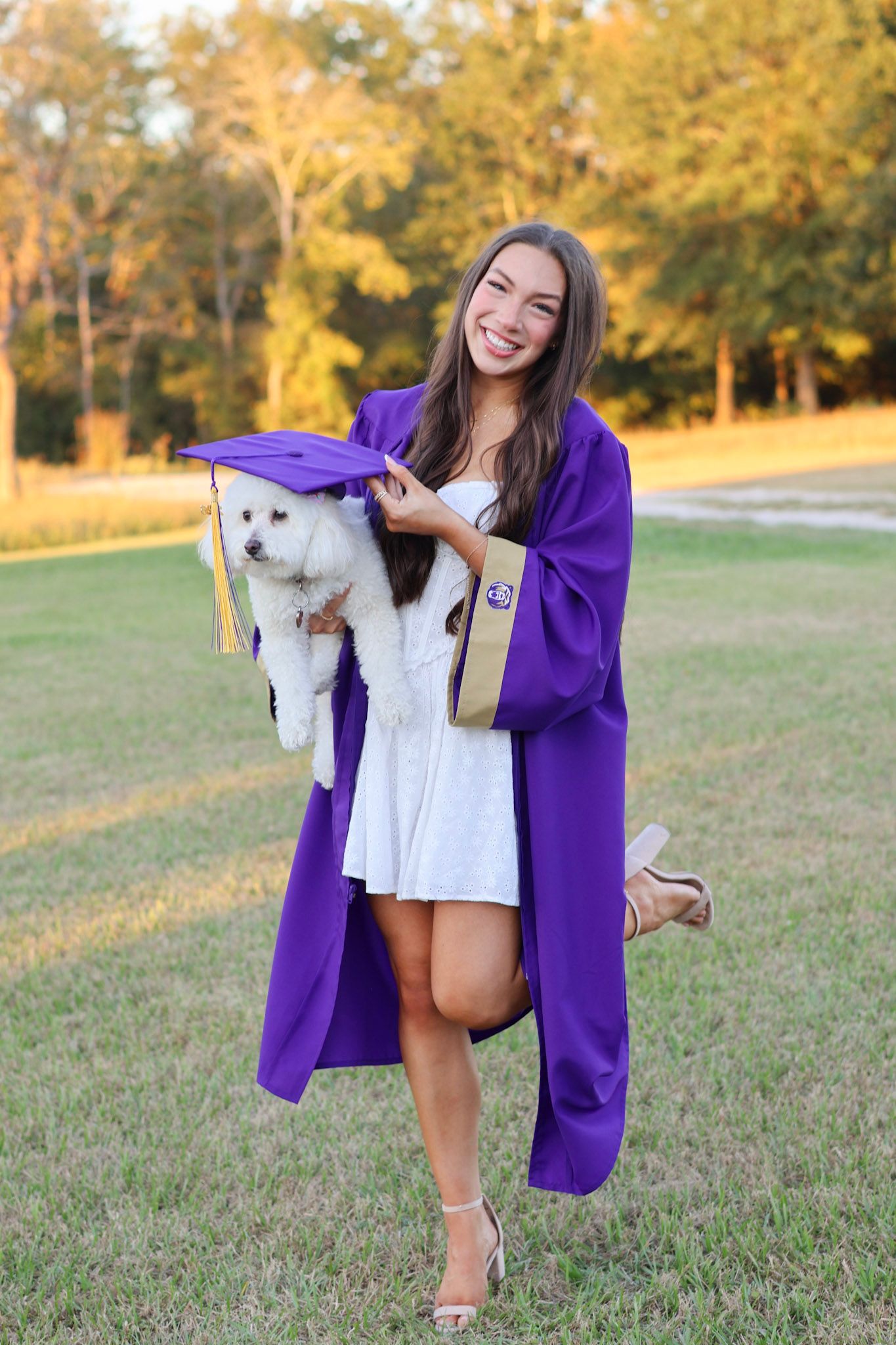 Smiling young woman in purple graduation gown and cap holding a white fluffy dog outdoors on green grass.