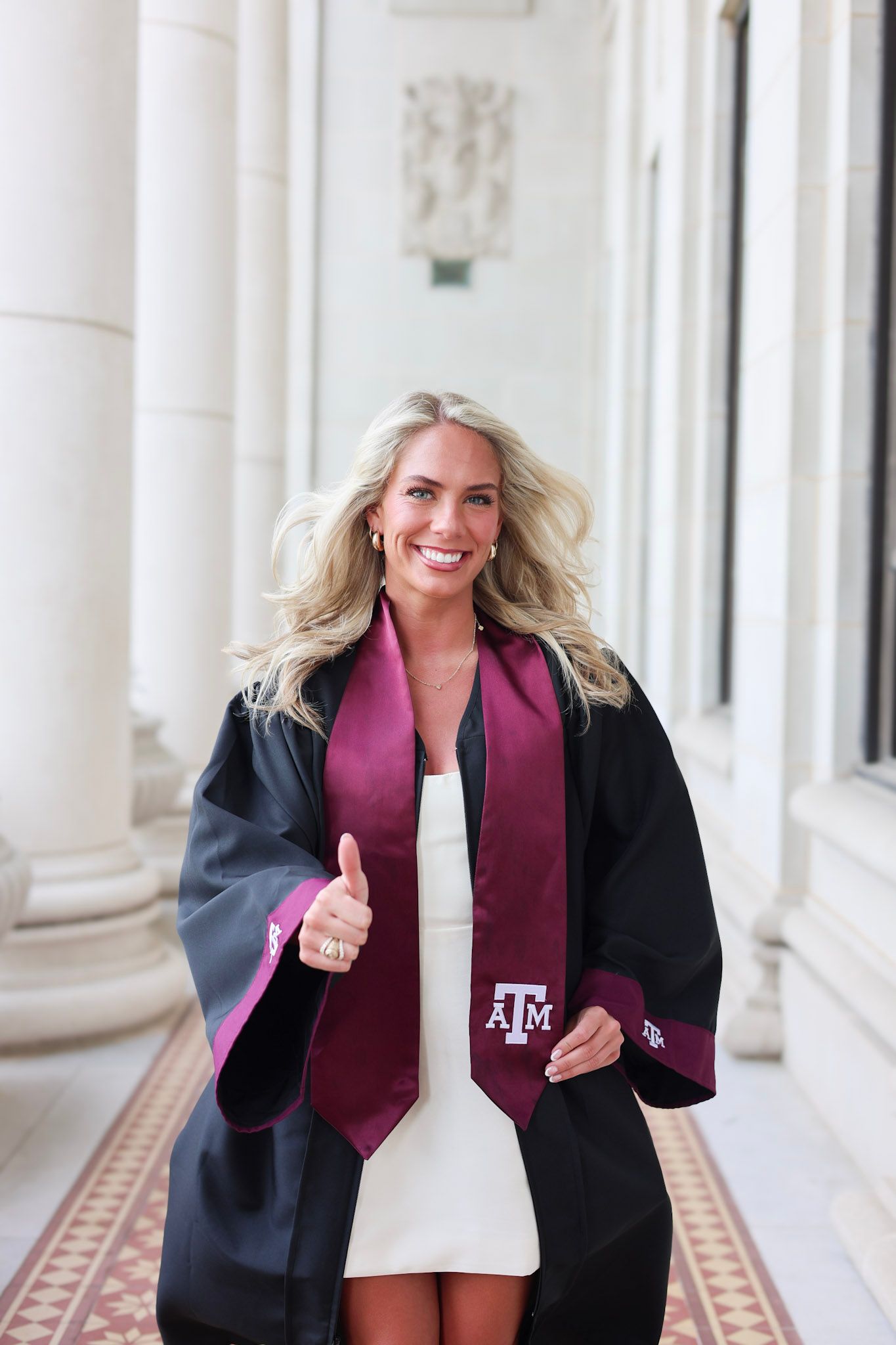 Smiling blonde woman in Texas A&M graduation gown and maroon stole giving thumbs up between white columns.