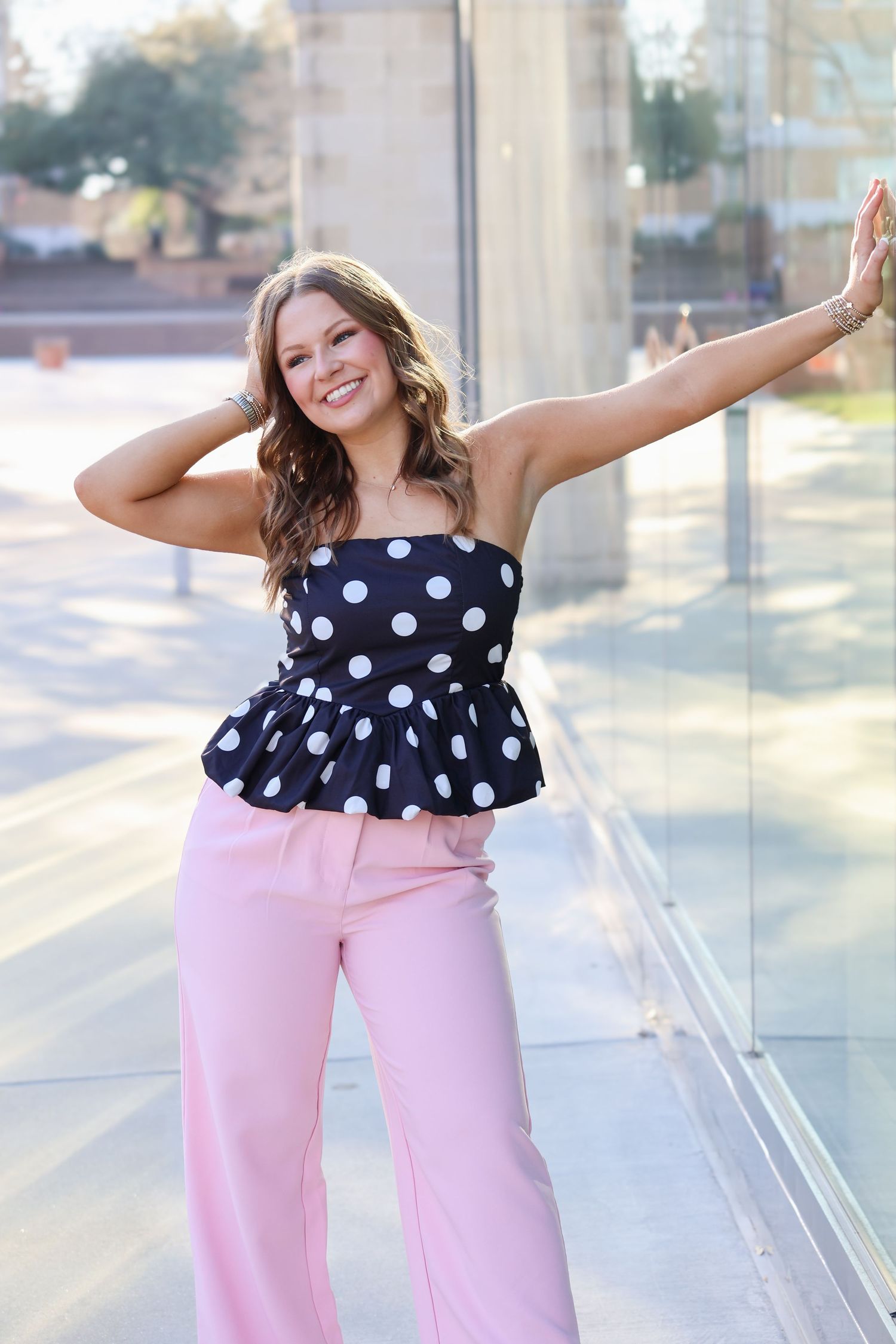 Woman in black polka dot peplum top and pink wide-leg pants posing outdoors near glass railing.