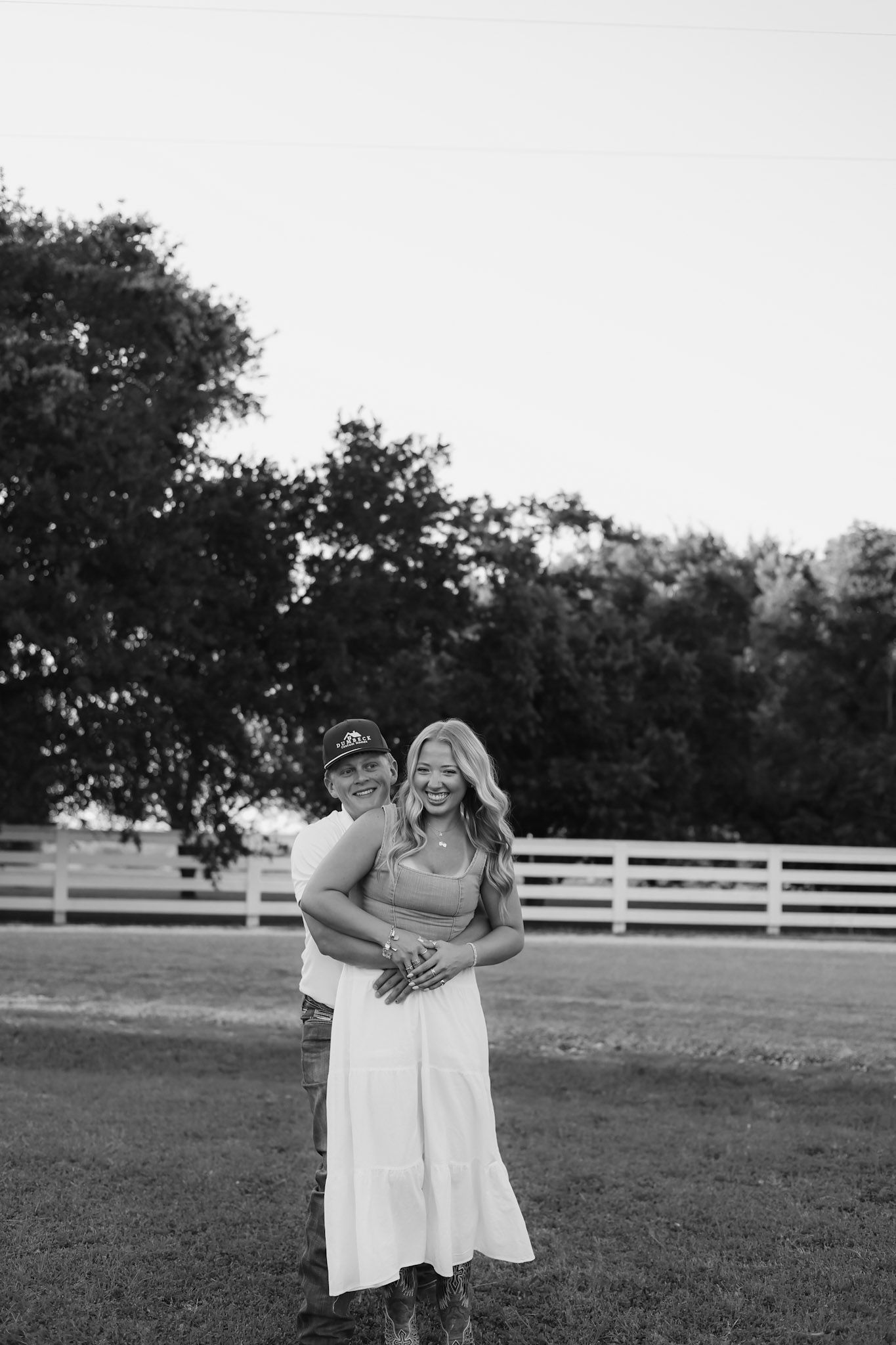 Black and white photo of a couple embracing in a field near a white fence and large trees.