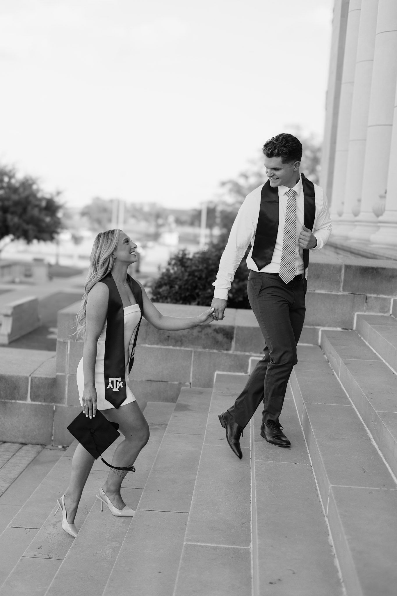 Black and white photo of Texas A&M graduate in cap and sash holding hands with suited man on stone steps.