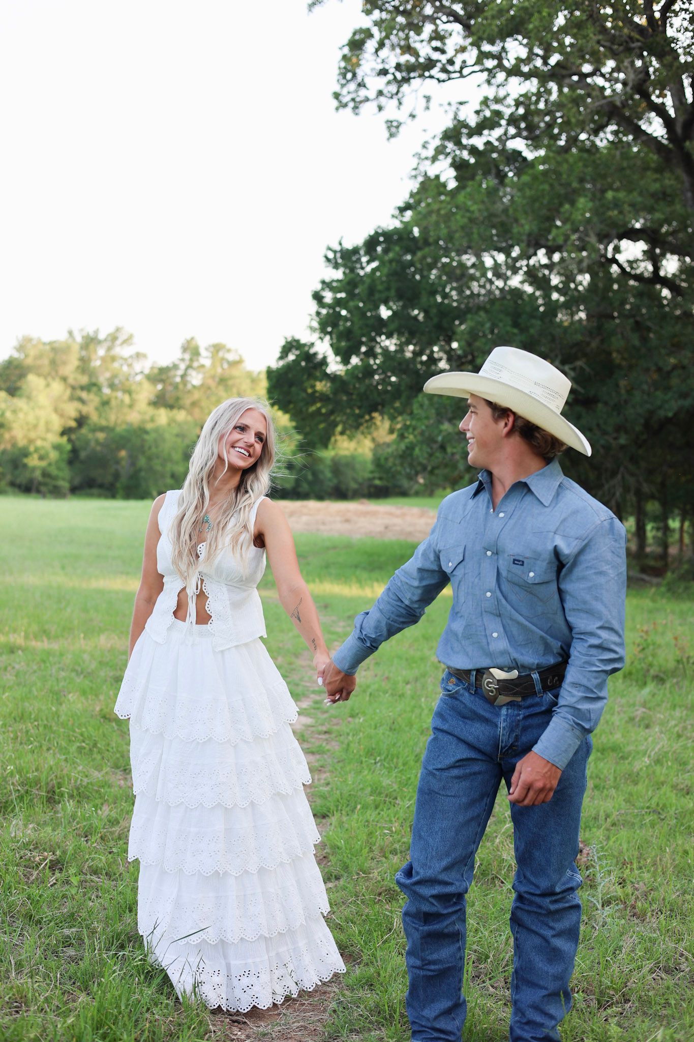 Smiling bride in white tiered dress holds hands with groom in cowboy hat and jeans in a green field.