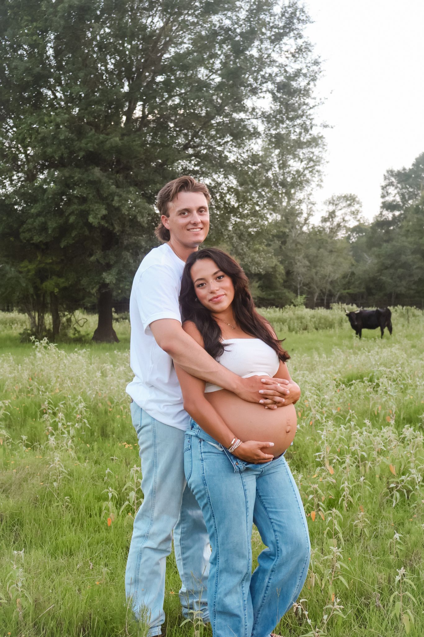 Couple embracing in casual outfits while standing in wildflower meadow with cattle in background.