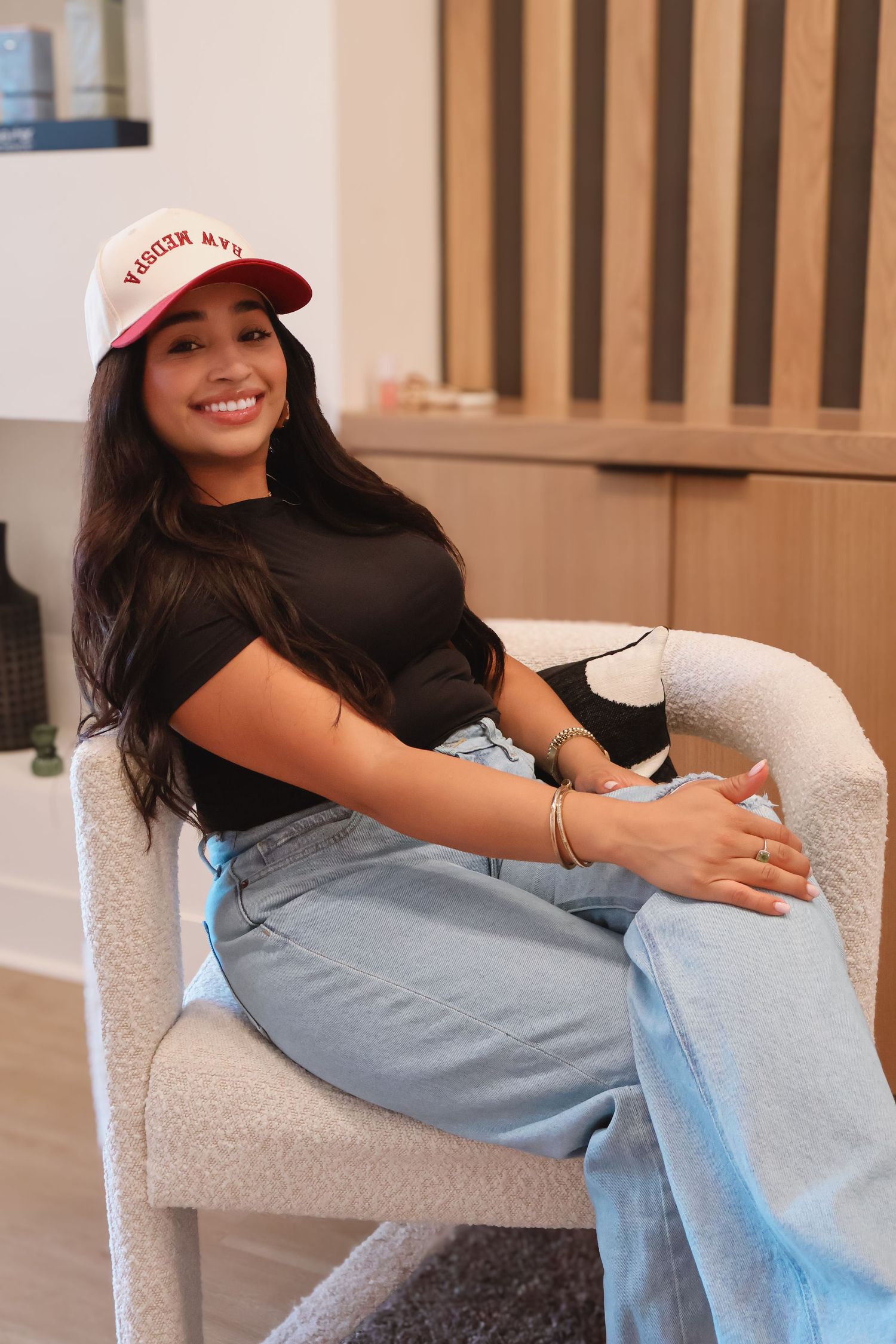 Woman in red cap and light denim jeans lounging on white armchair in casual photoshoot sequence.