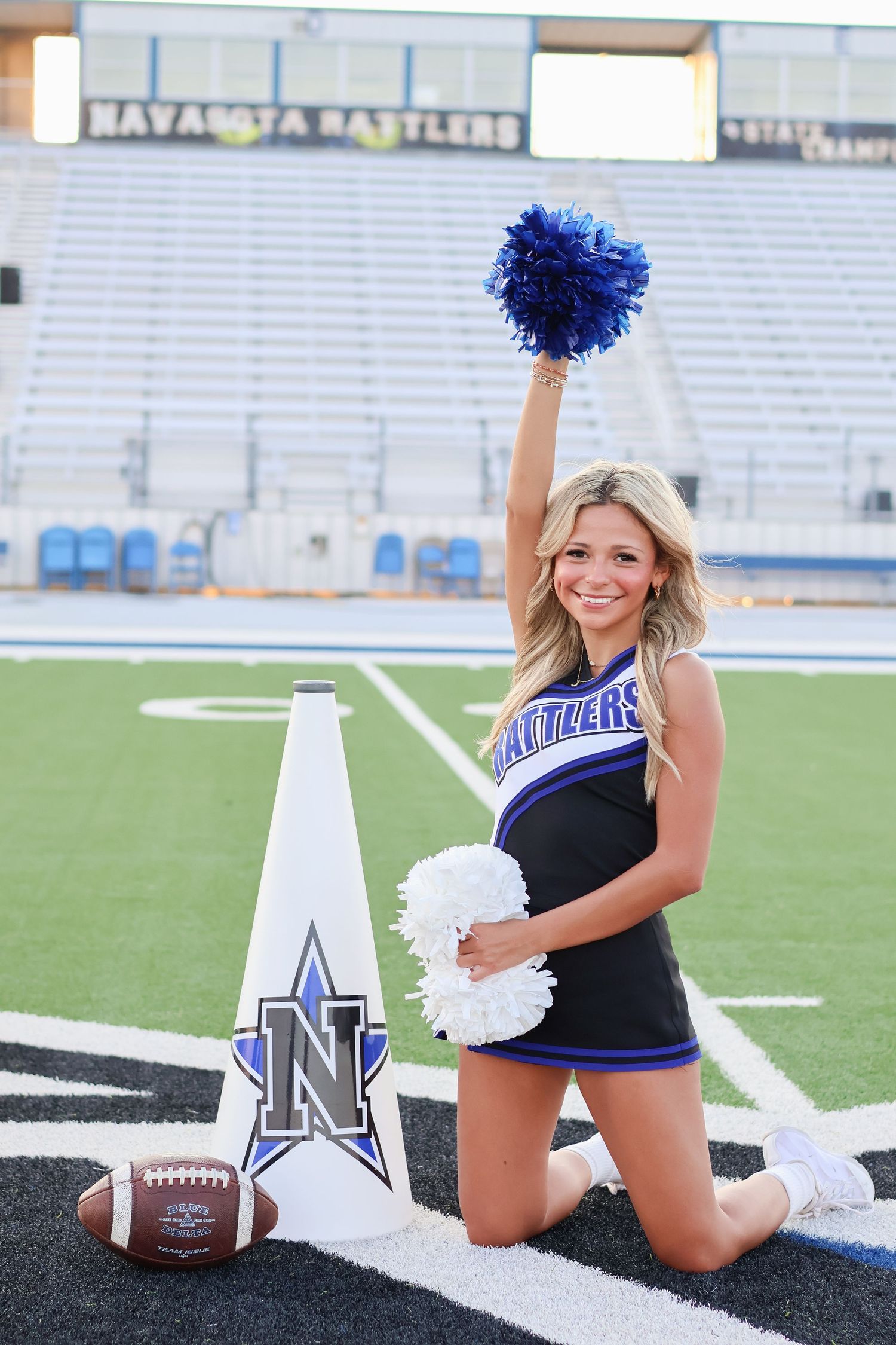 Cheerleader with pom poms poses with football on sports field near white goalpost marker.