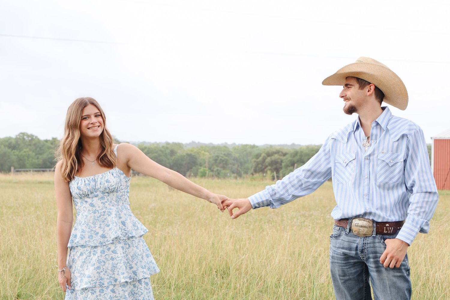 Couple holding hands in field wearing western attire with cowboy hat and floral dress.