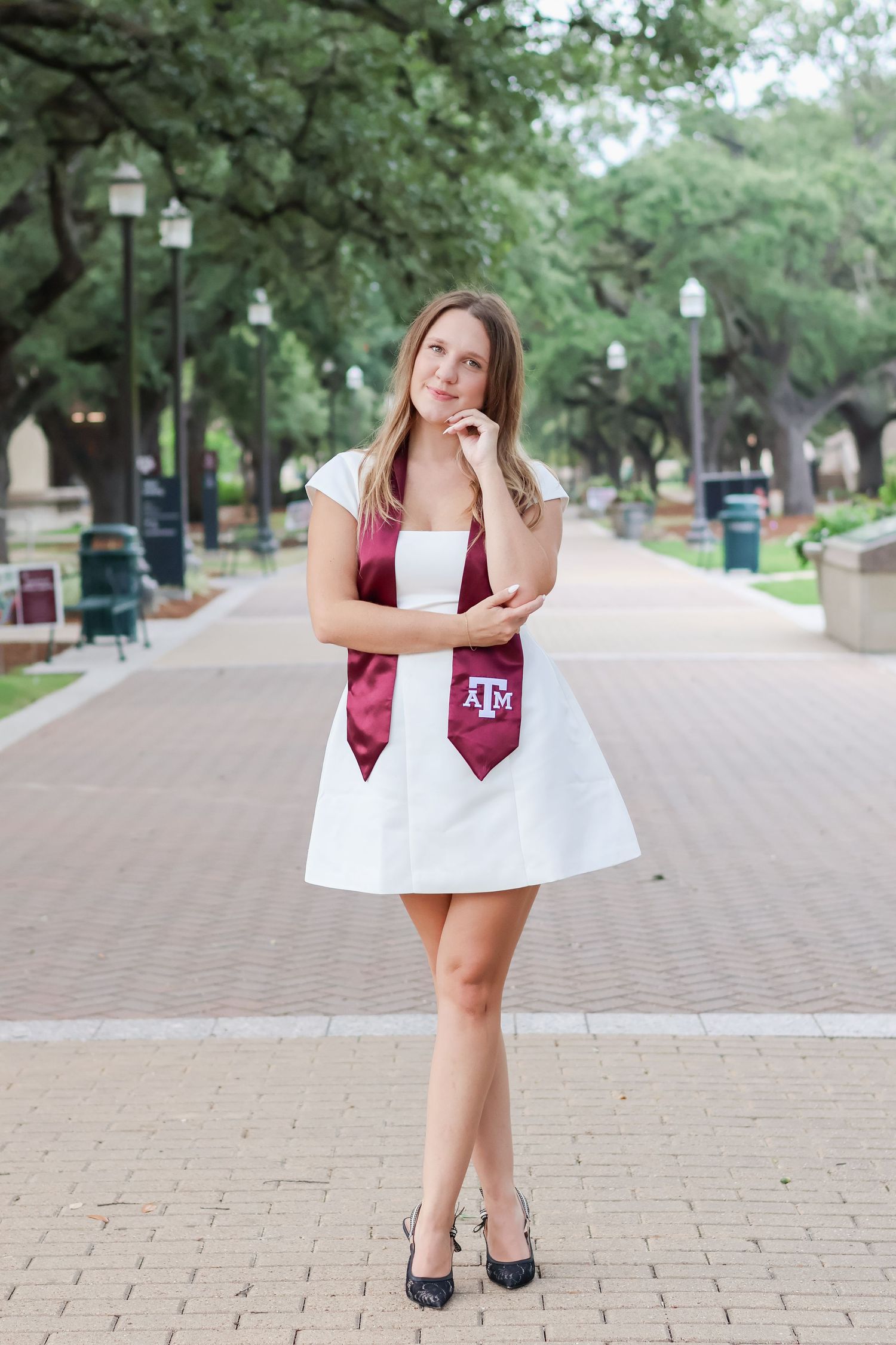 A graduate in a white dress and maroon Texas A&M stole poses thoughtfully on a tree-lined brick pathway with lamp posts.
