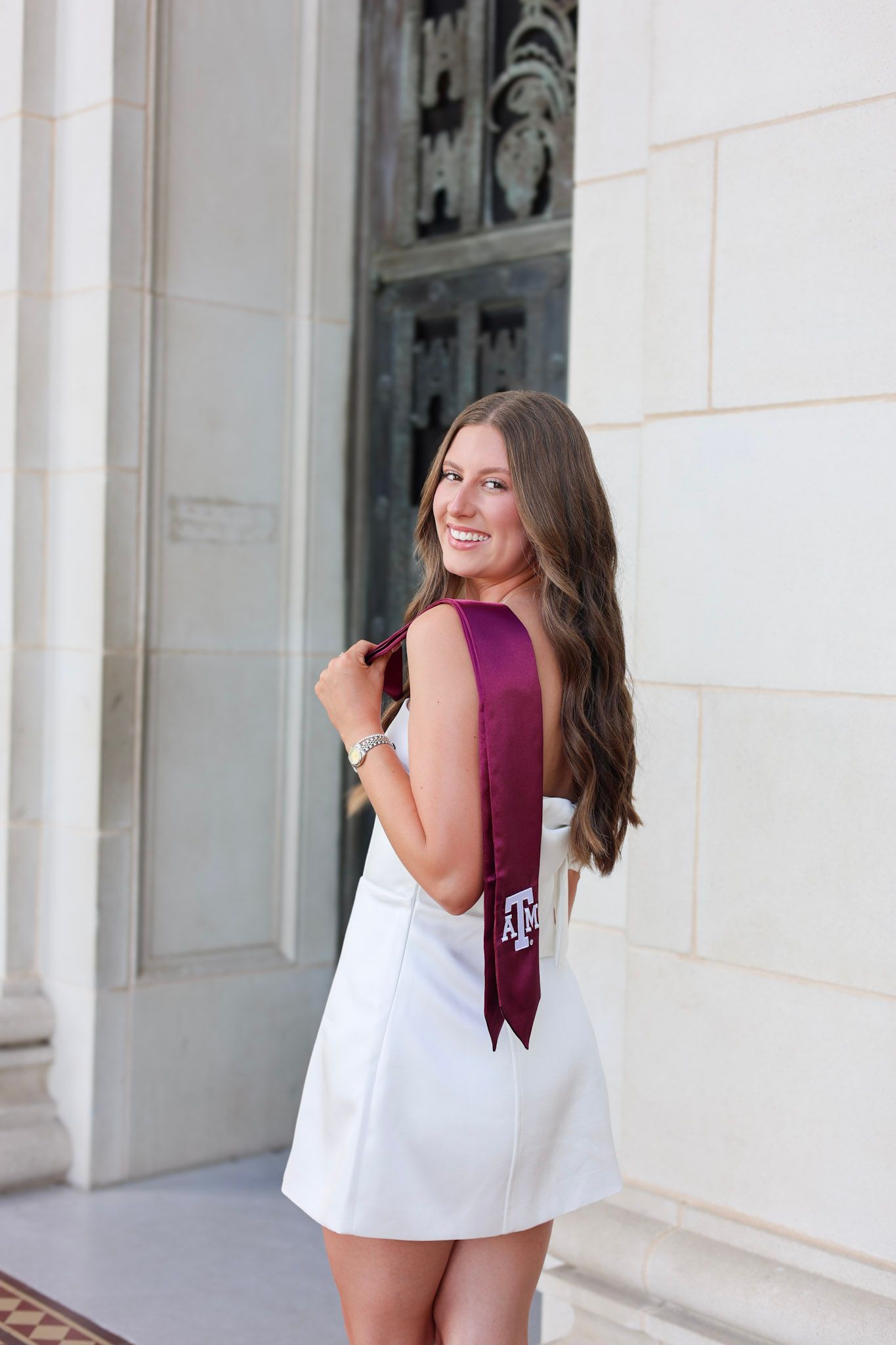 Smiling brunette woman in white dress with maroon Texas A&M graduation sash poses at stone building.
