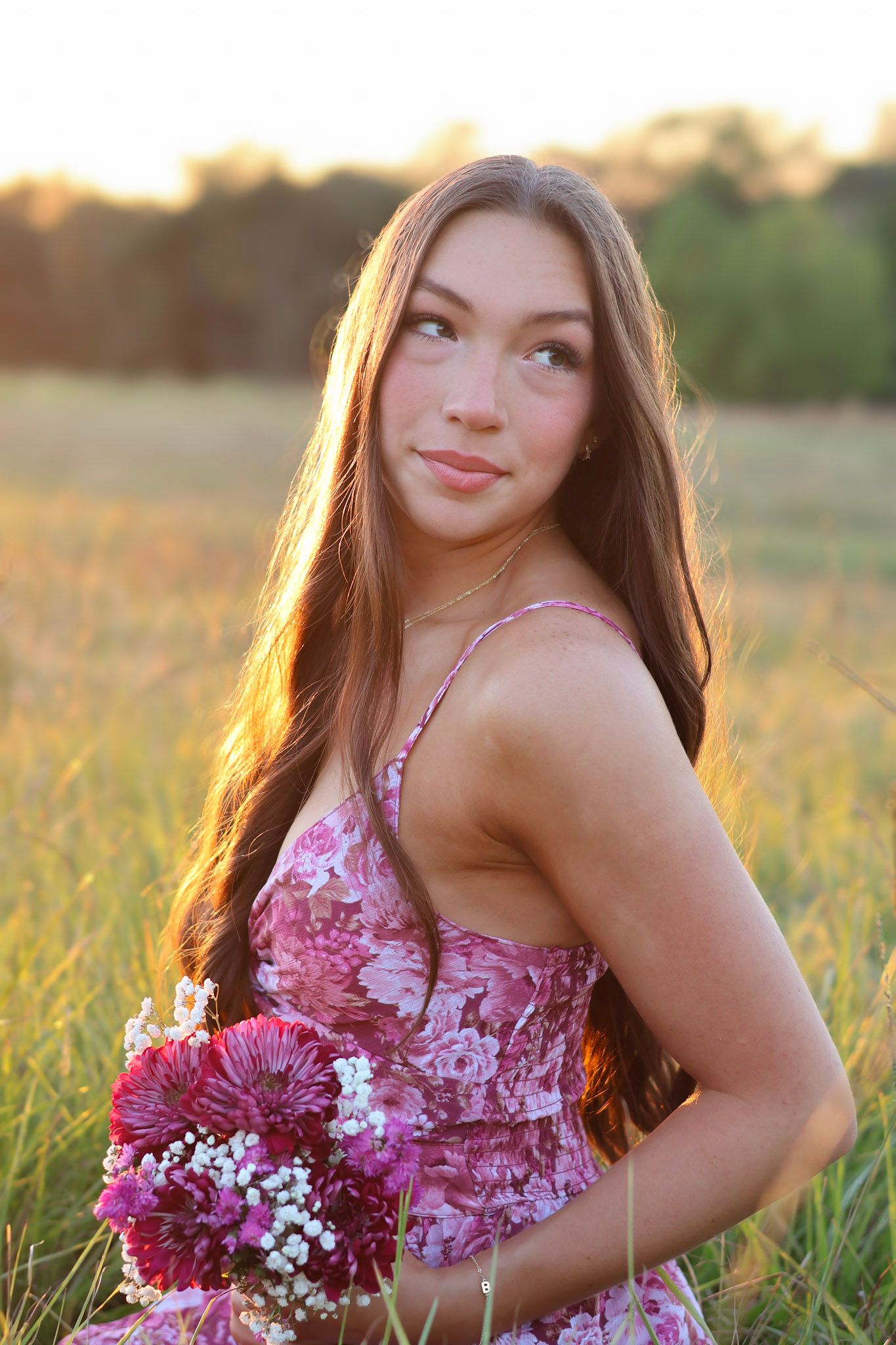 Young woman in floral dress holding pink flowers poses in golden sunlit field with long brown hair.