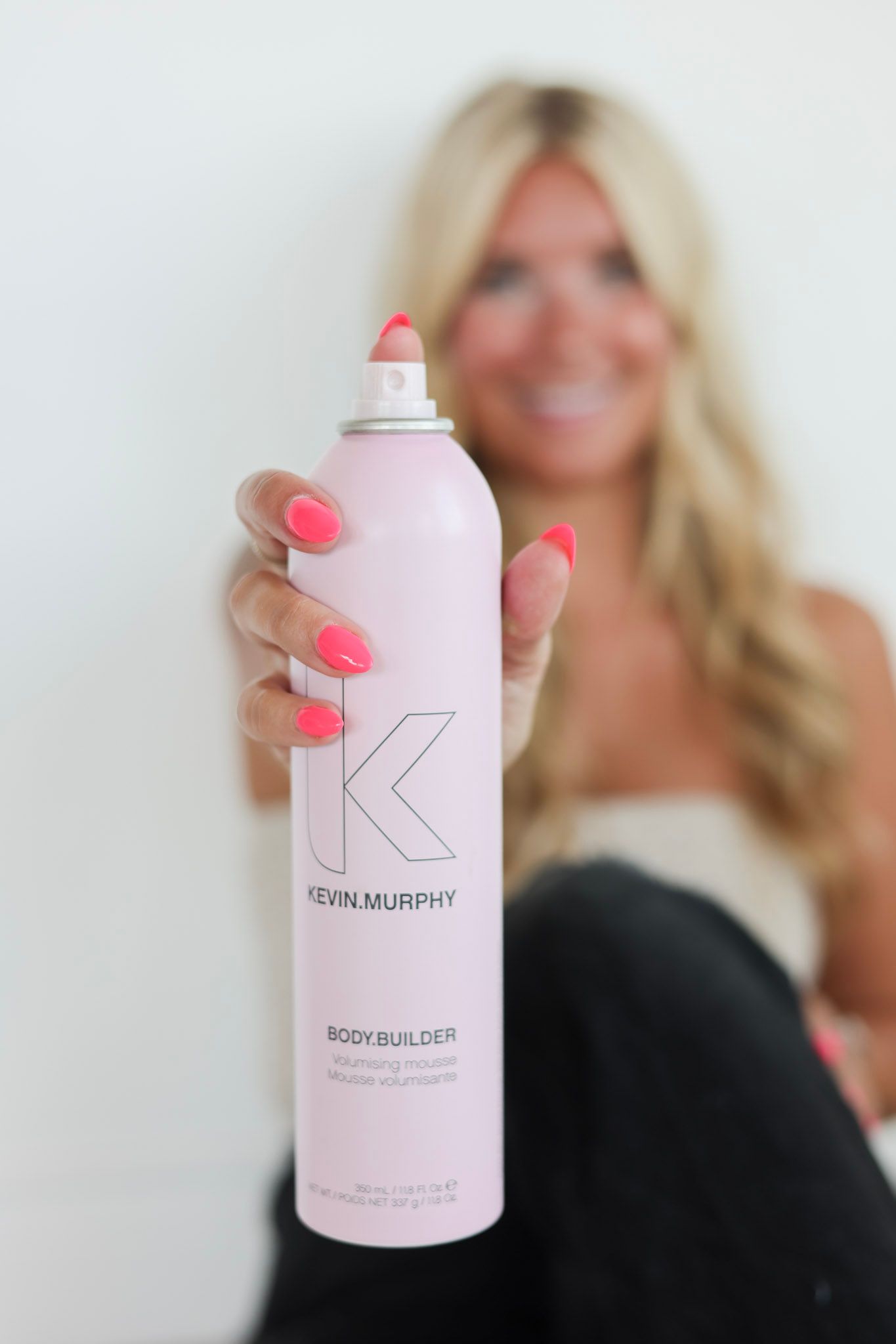 Blonde woman holding Kevin Murphy Body Builder volumizing mousse spray can with bright pink nails.