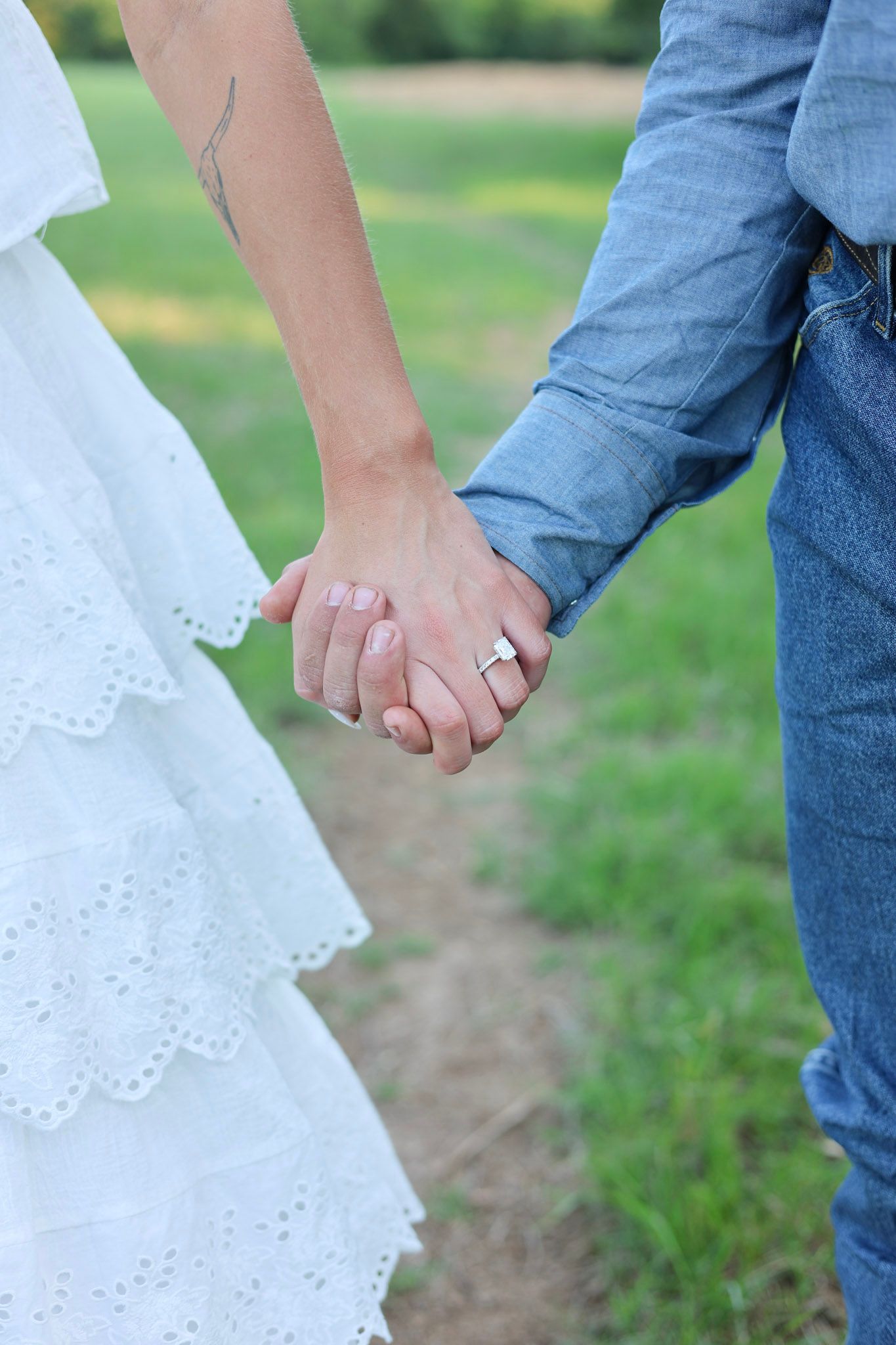 Newlywed couple holding hands in a green field, bride in white lace dress with wedding ring visible.