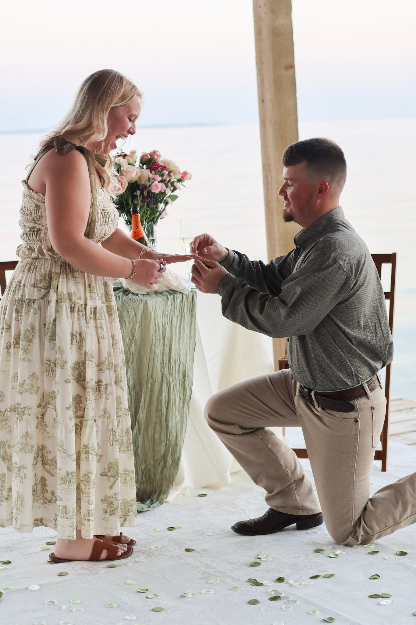 Man kneeling on one knee proposing to woman with flowers by the ocean at sunset.