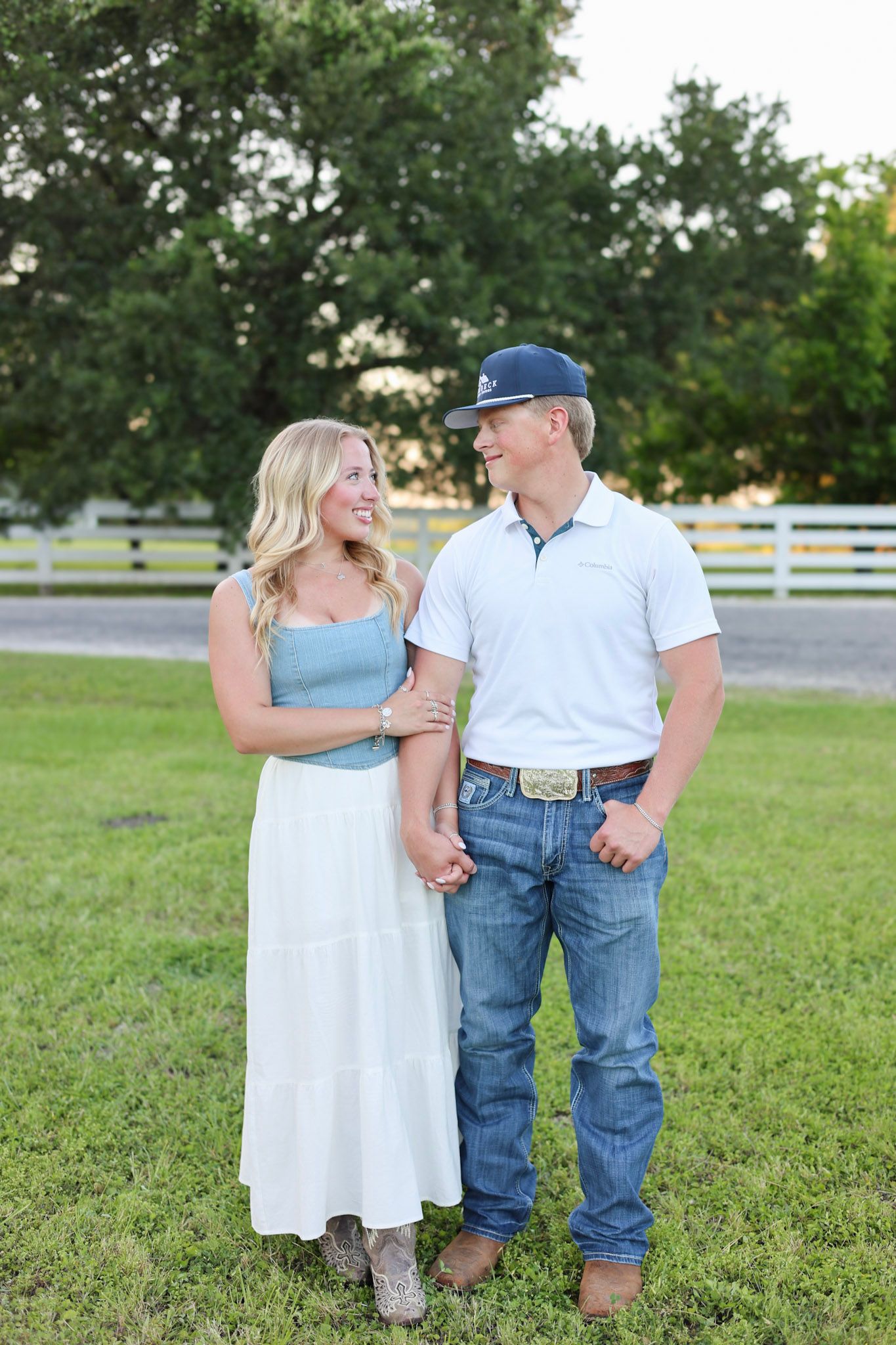 Smiling couple holding hands in a sunny farm field, woman in white skirt and man in jeans and cap.