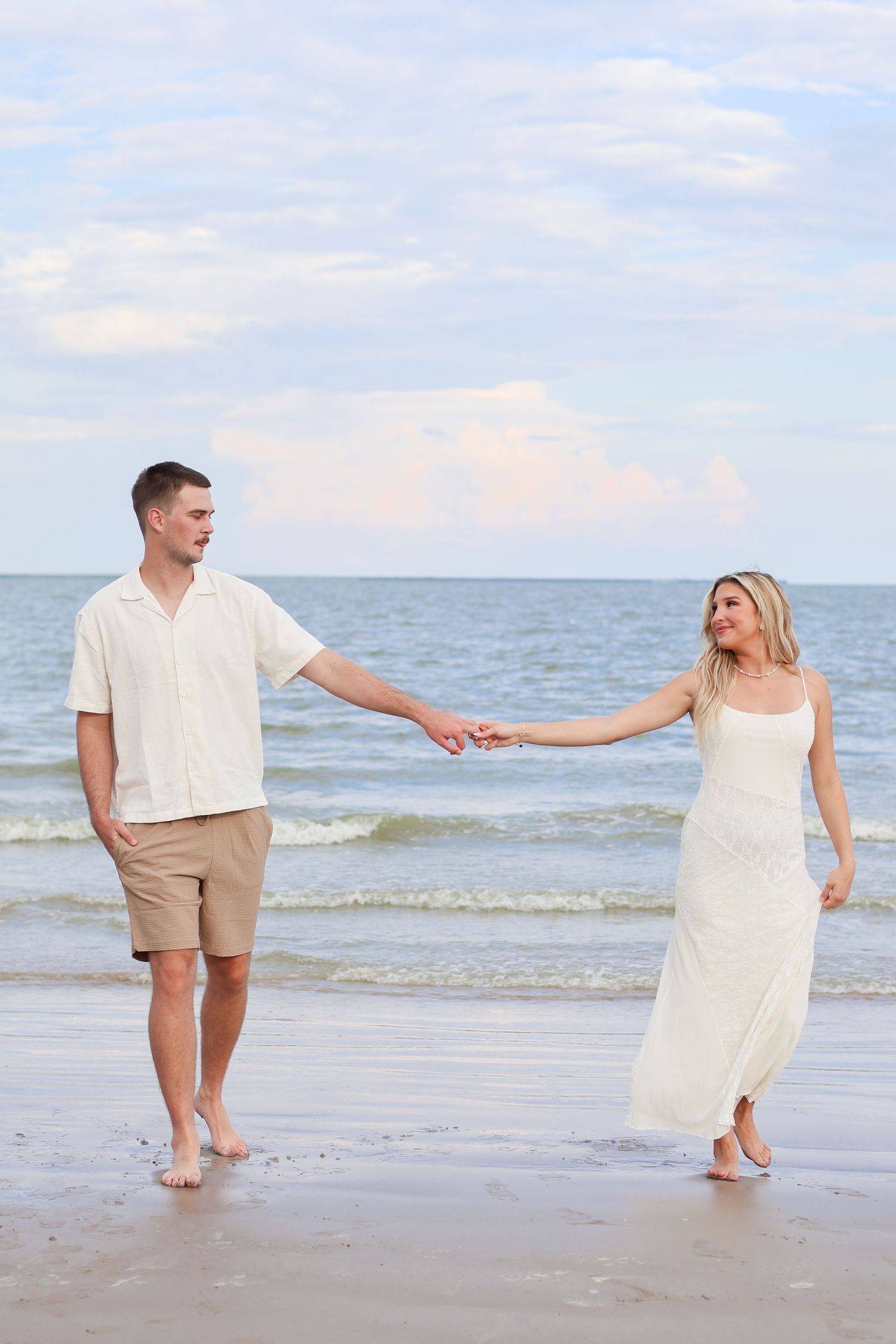 A couple in beachwear holding hands and walking along the shoreline at sunset with ocean waves in the background.