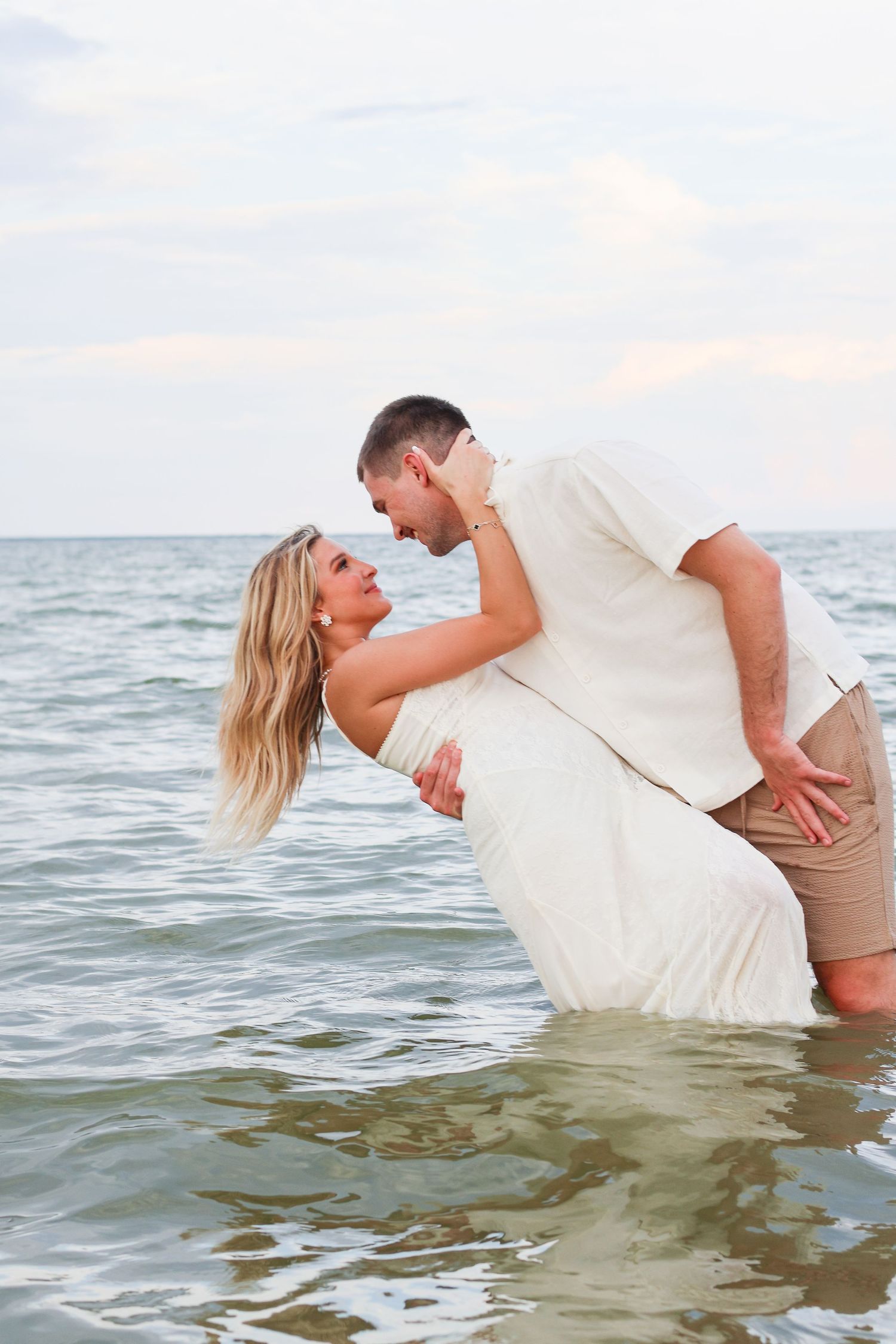 A romantic couple in white clothing shares a tender moment while standing in ocean waters at sunset.