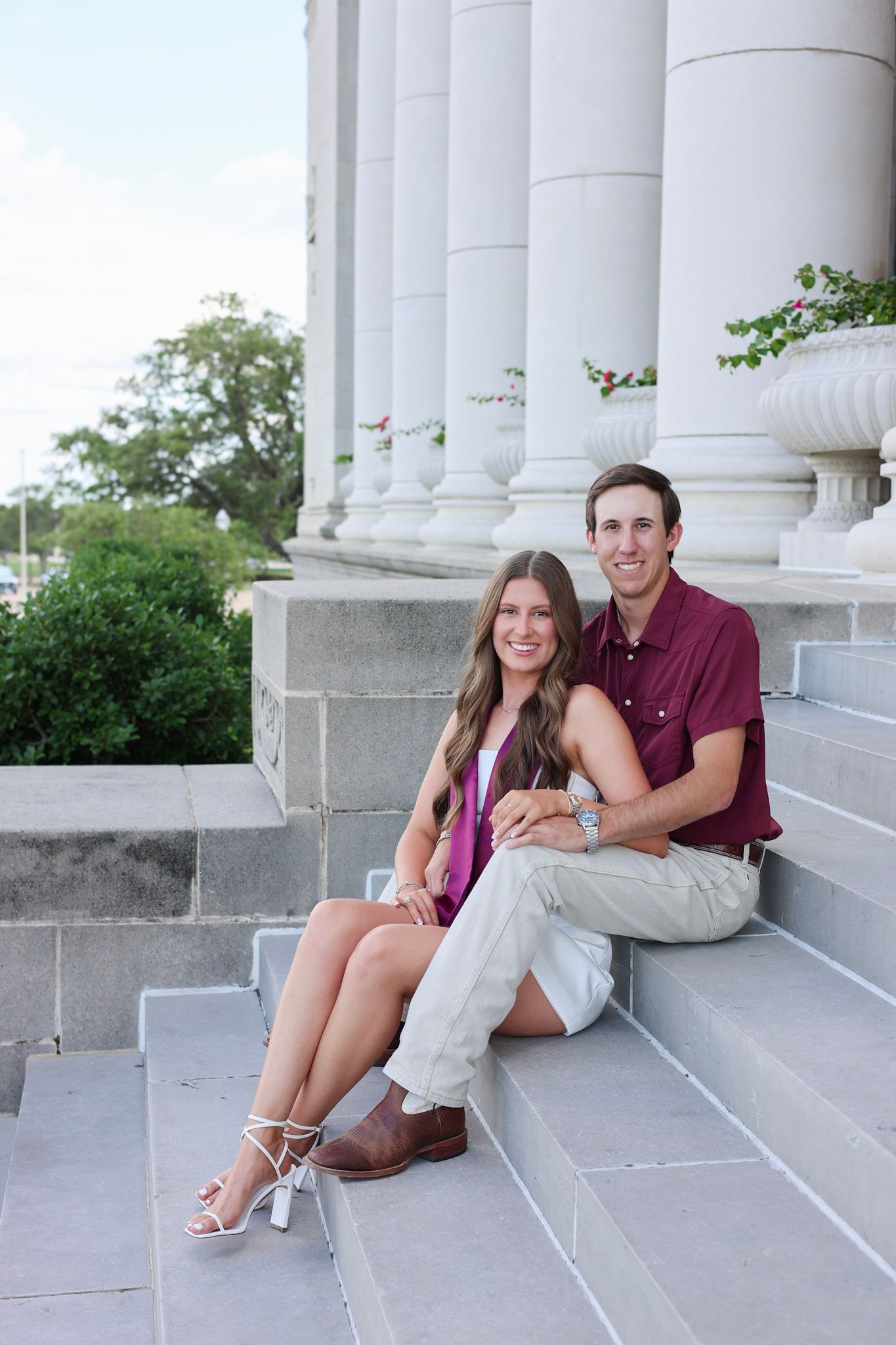 Couple sitting on stone steps of a classical building with white columns, smiling for engagement photos.