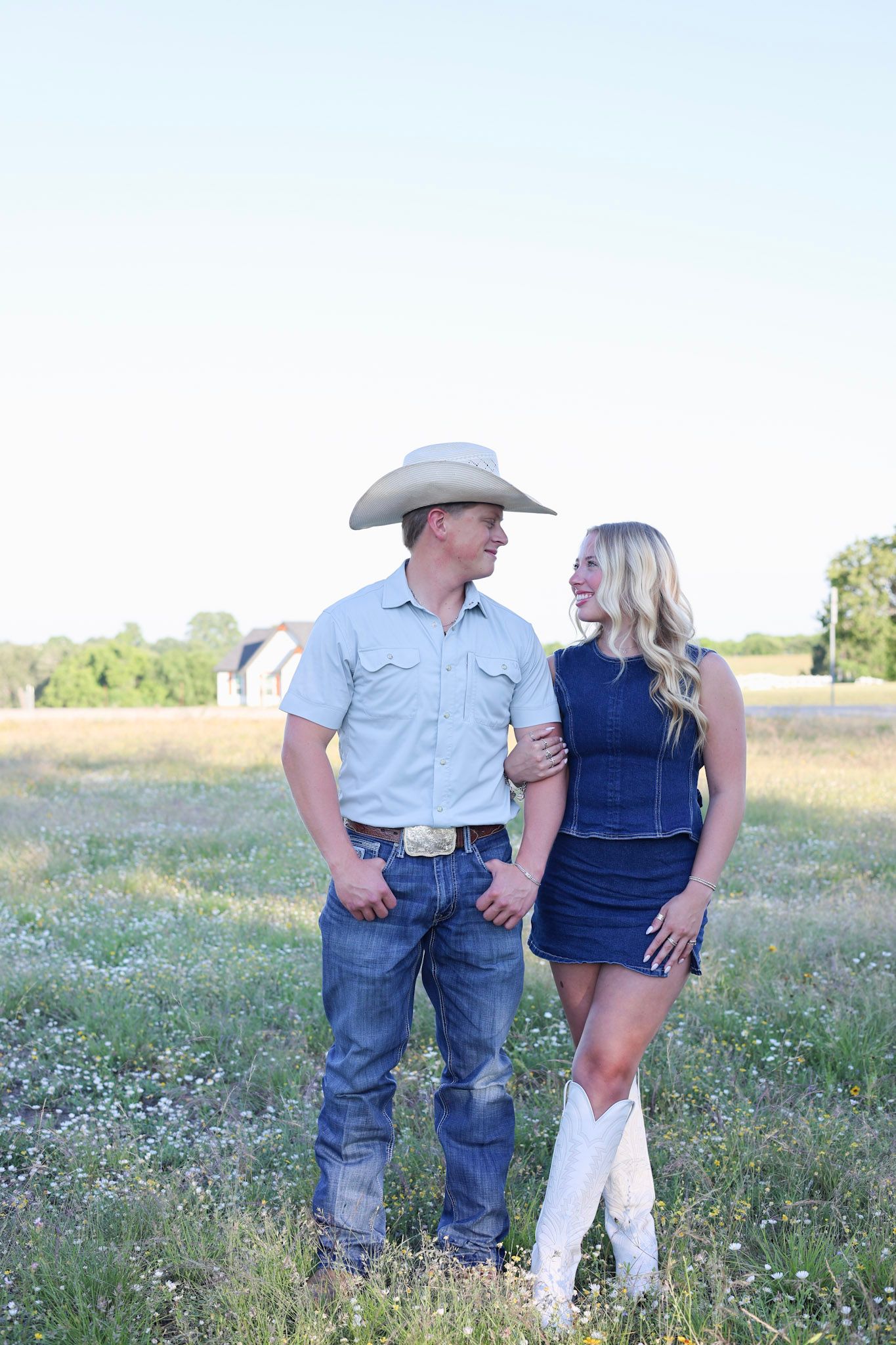 A couple in Western attire stands in a wildflower field, man in cowboy hat and jeans, woman in denim dress and white boots.