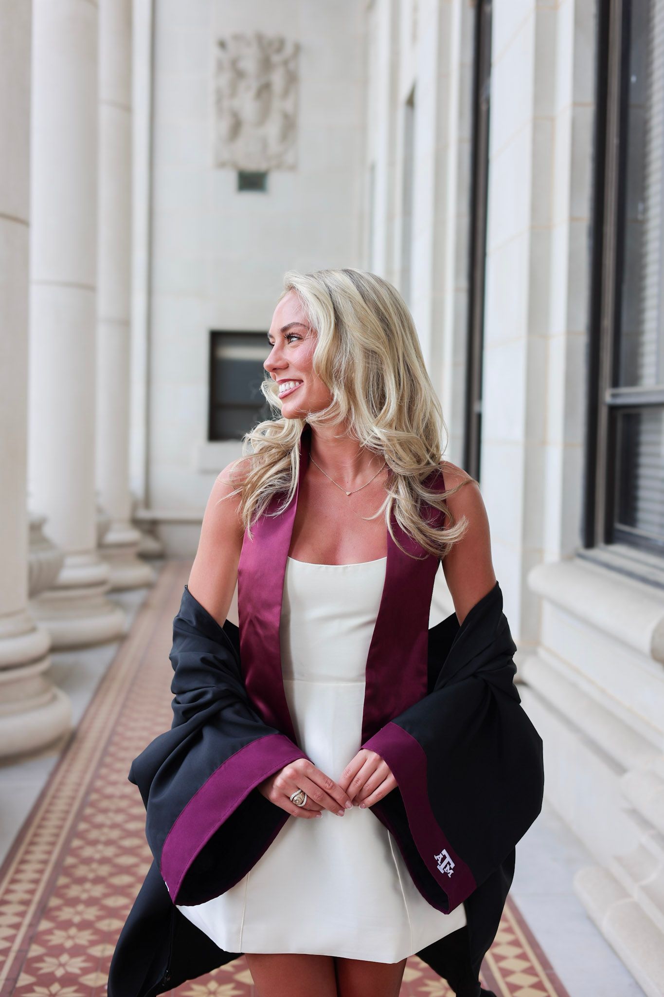 Smiling blonde woman in white dress with maroon Texas A&M graduation stole and black gown between columns.