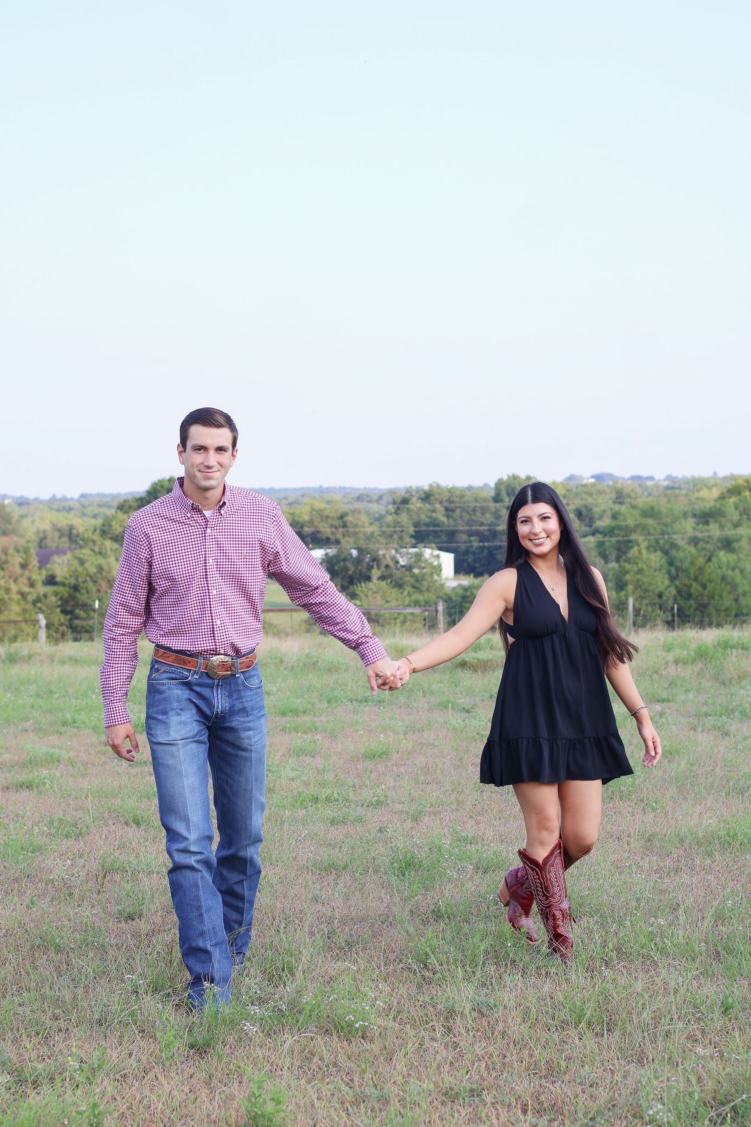 A couple holding hands while walking through a grassy field, with her wearing a black dress and cowboy boots.