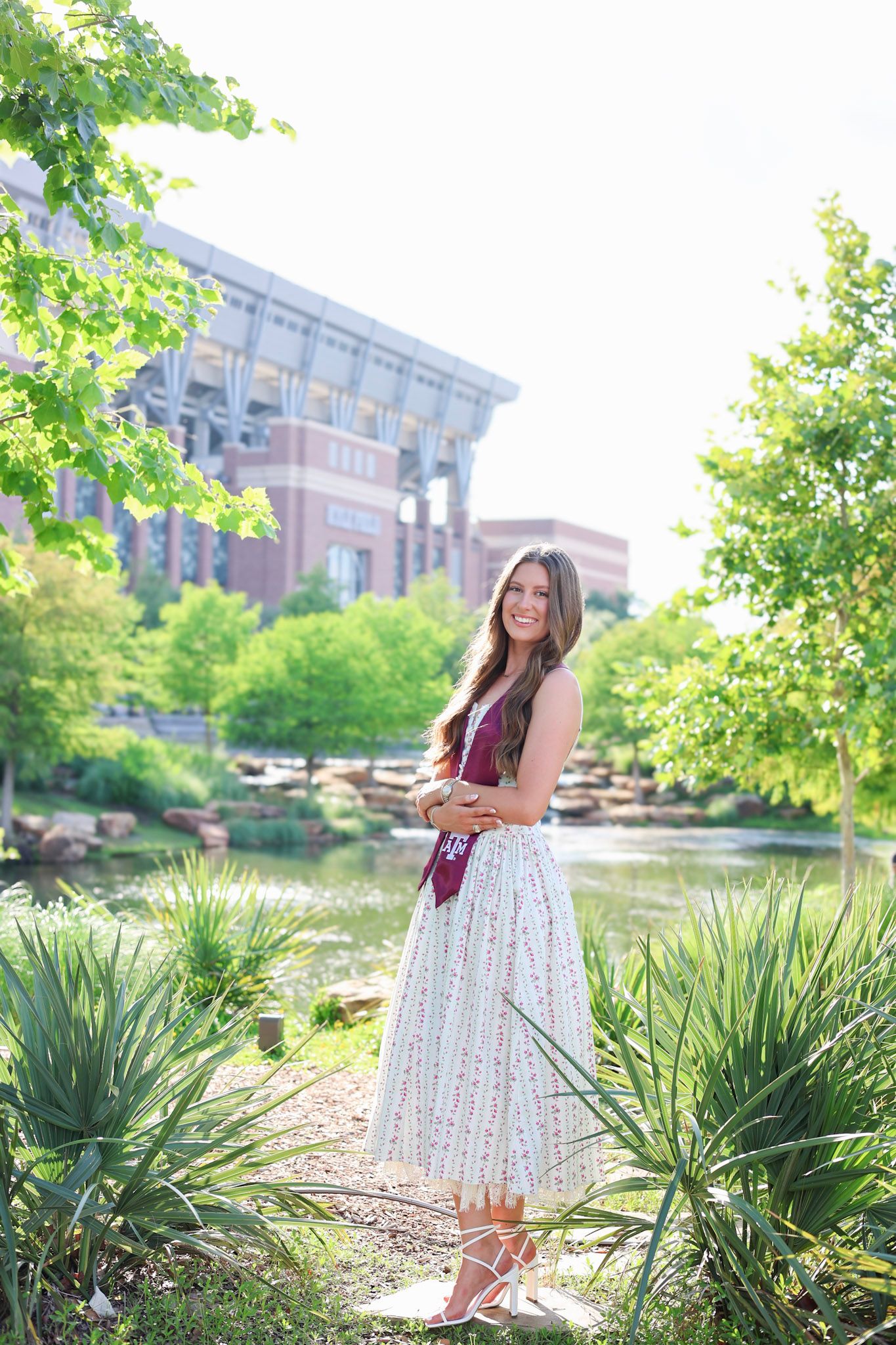 Young woman in floral skirt and maroon top poses in a lush garden near a brick building on a sunny day.