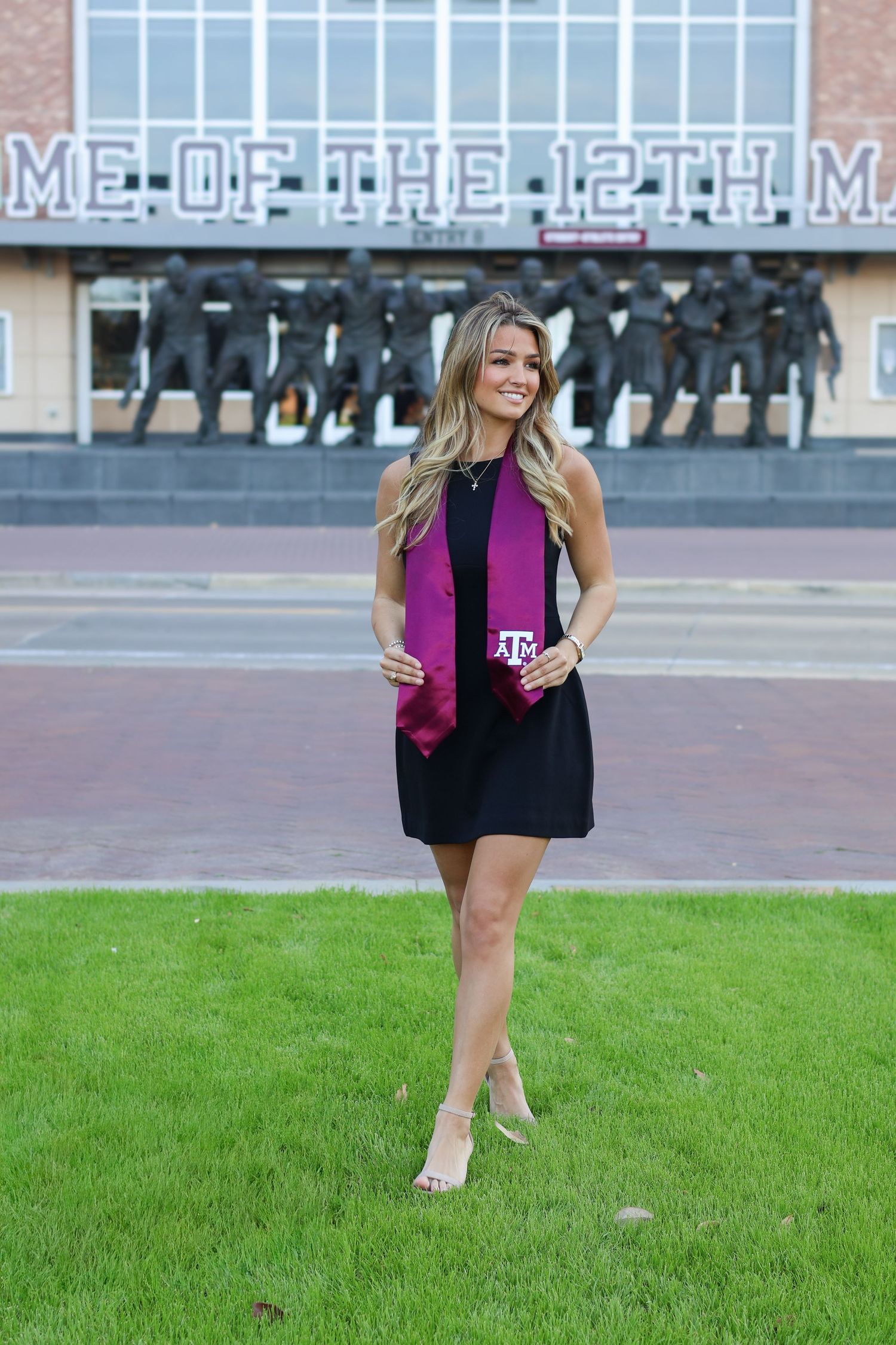 Young woman in black dress with maroon Texas A&M graduation stole posing outside Kyle Field stadium.