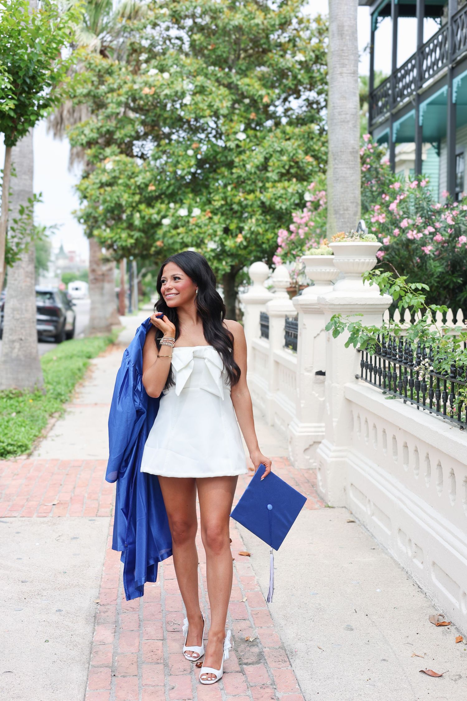 Graduate in white bow dress carries blue cap and gown on a sunny brick sidewalk.