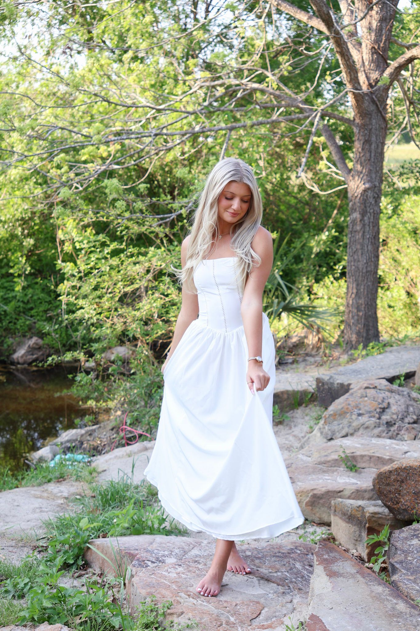 Blonde woman in white maxi dress standing barefoot on rocks near creek in nature.