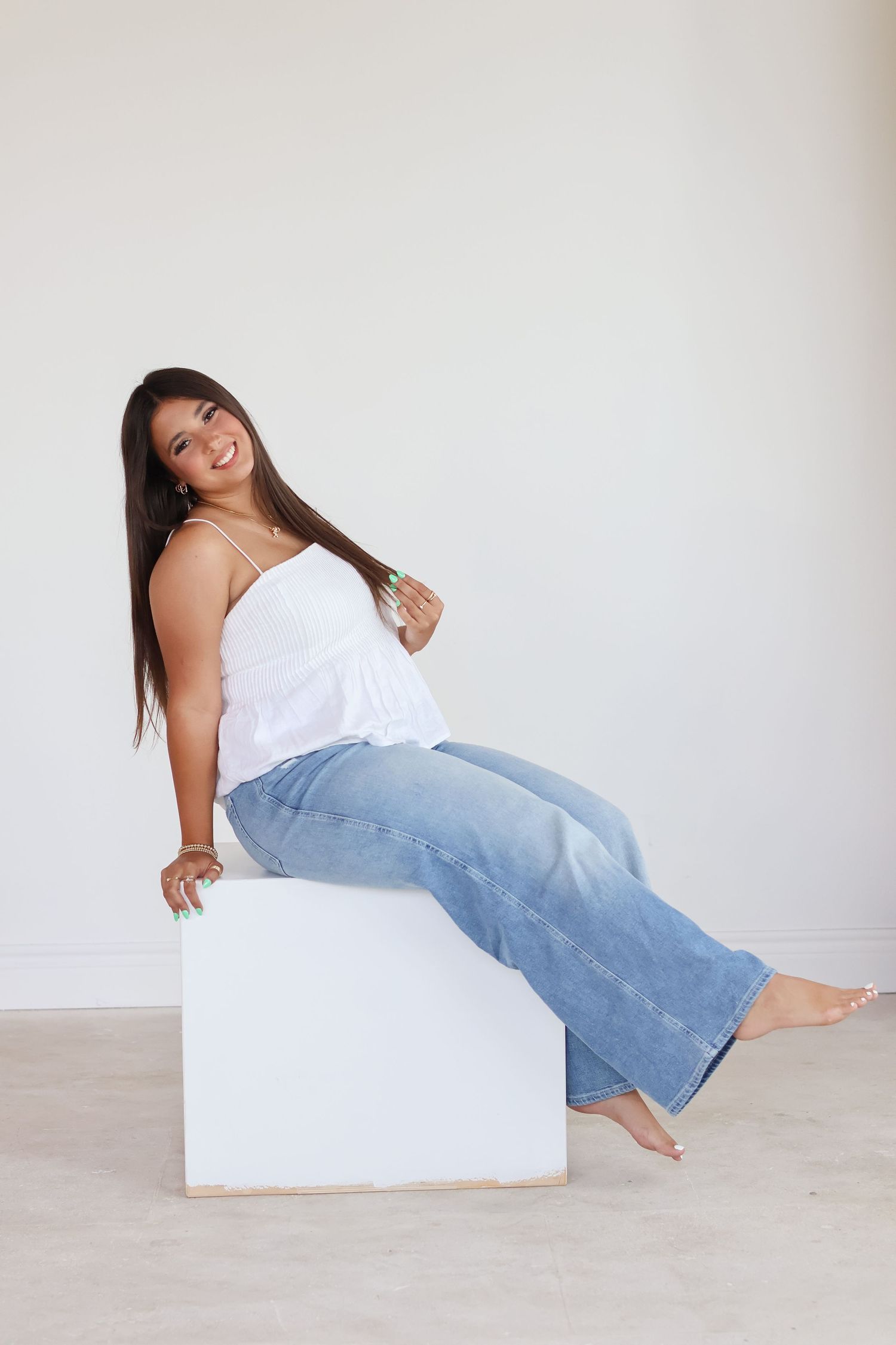 A person in a white top and light blue jeans poses playfully on a white platform against a neutral background.