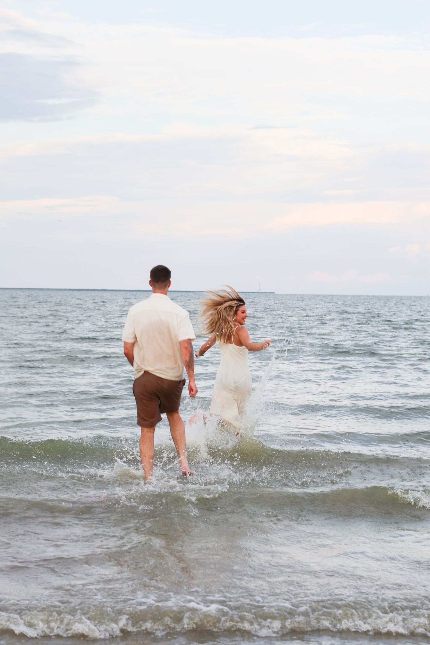 A couple in white and brown attire playfully splashes through ocean waves at sunset.