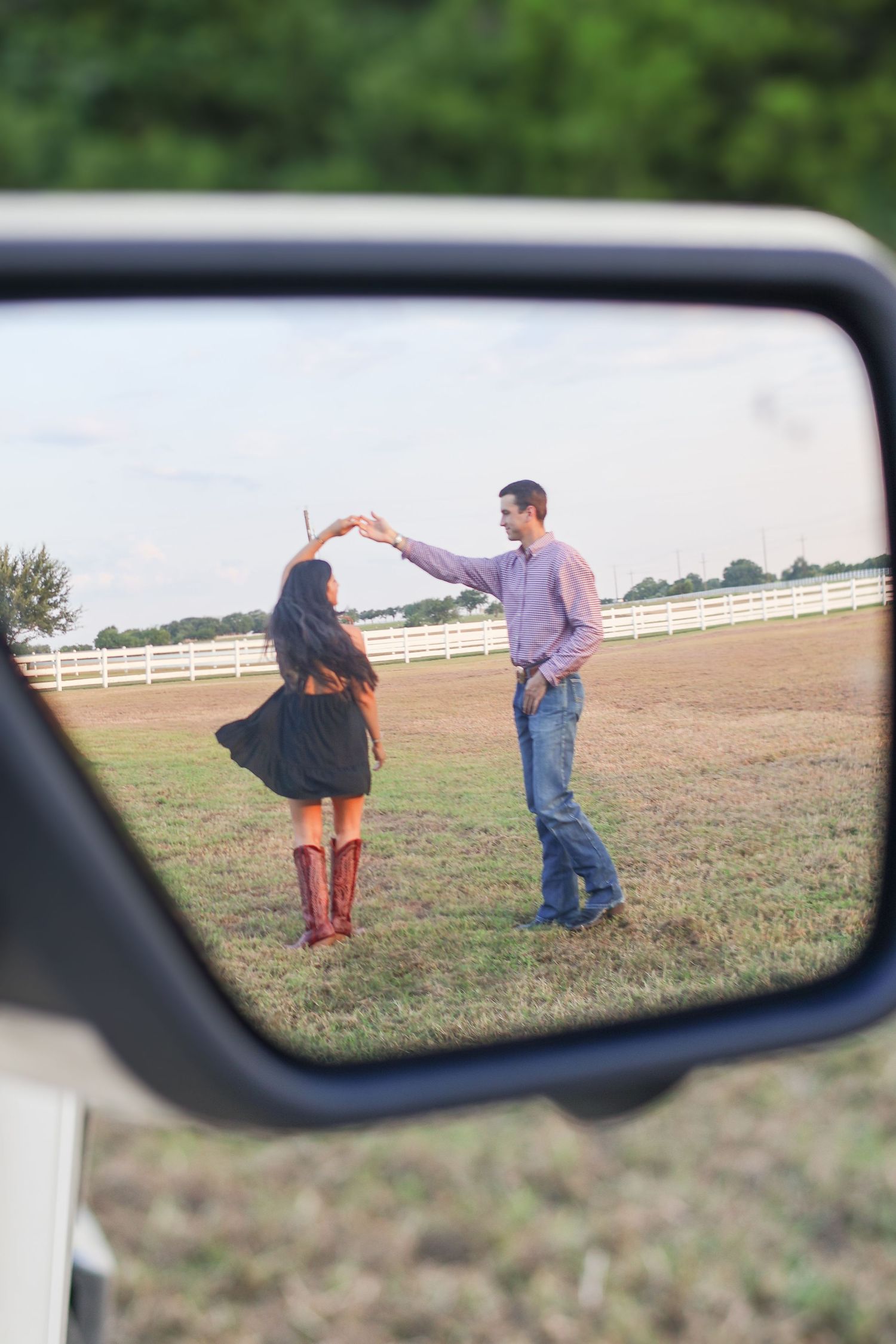 A couple is captured in a car's rearview mirror as they stand together in a field with a white fence in the background.