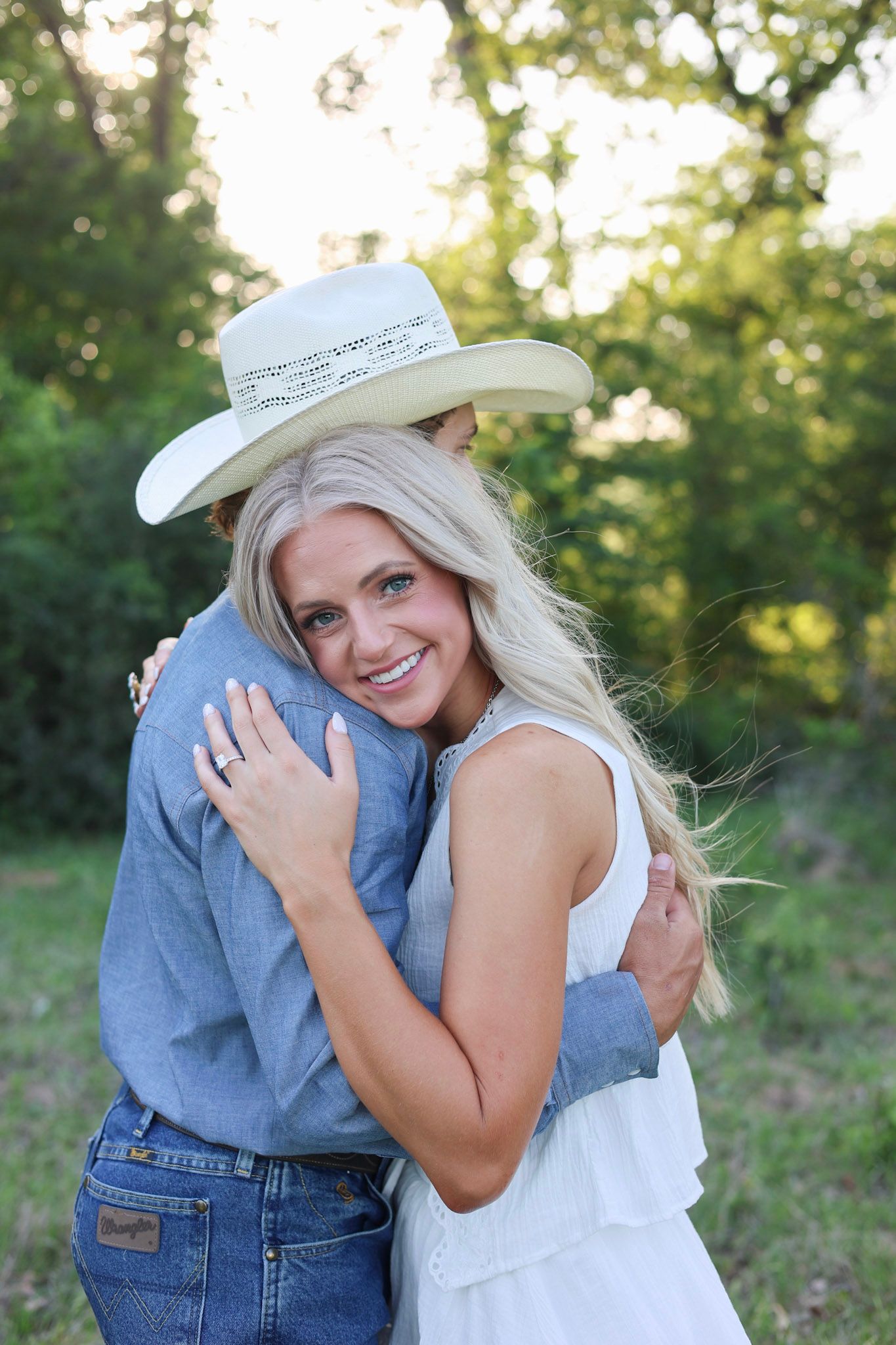 A blonde woman in a white dress hugging a man in a cowboy hat and blue shirt in a green outdoor setting.